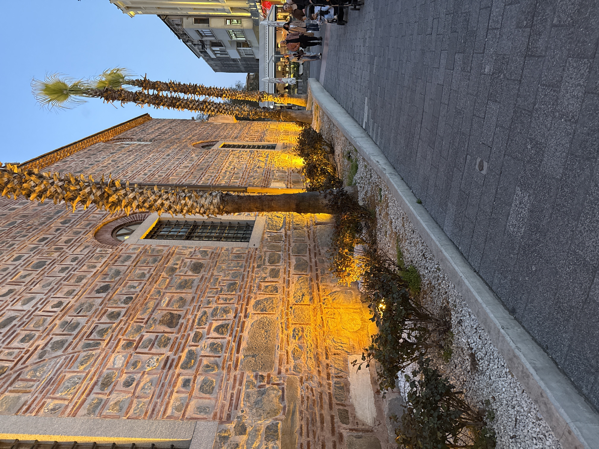 A long shot depicts an outdoor scene, likely in Plovdiv, Bulgaria, captured during the evening. The image focuses on the side of a stone building, possibly a historical structure, featuring a distinct pattern of stones and brick. Illumination from spotlights casts a warm yellow glow on the building's facade. Palm trees line the walkway adjacent to the building. A raised bed separates the building from the stone-paved pedestrian walkway. People are visible in the background, suggesting an active urban setting.