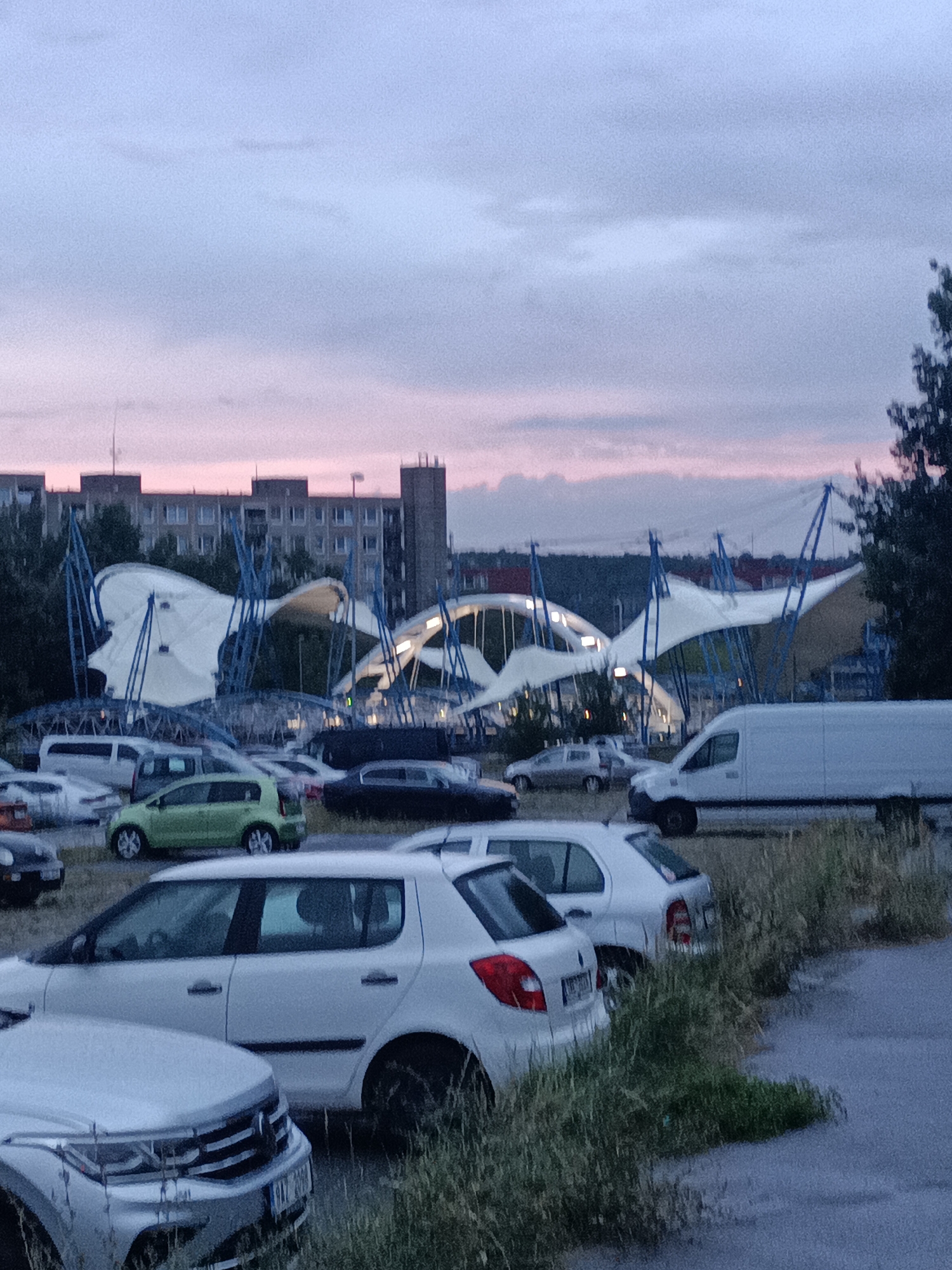 The image depicts an outdoor scene at dusk, featuring a parking lot filled with various vehicles, including cars and a white van. In the background, a modern architectural structure with white fabric canopies supported by blue poles is visible, alongside a multi-story building. The sky shows a gradient of colors, transitioning from light blue at the top to pink and purple near the horizon, indicating twilight. The foreground is dominated by parked cars and overgrown grass.
While the image contains elements of a parking lot and modern architecture, it does not provide sufficient visual cues to definitively identify the specific location within Prague, Czechia.