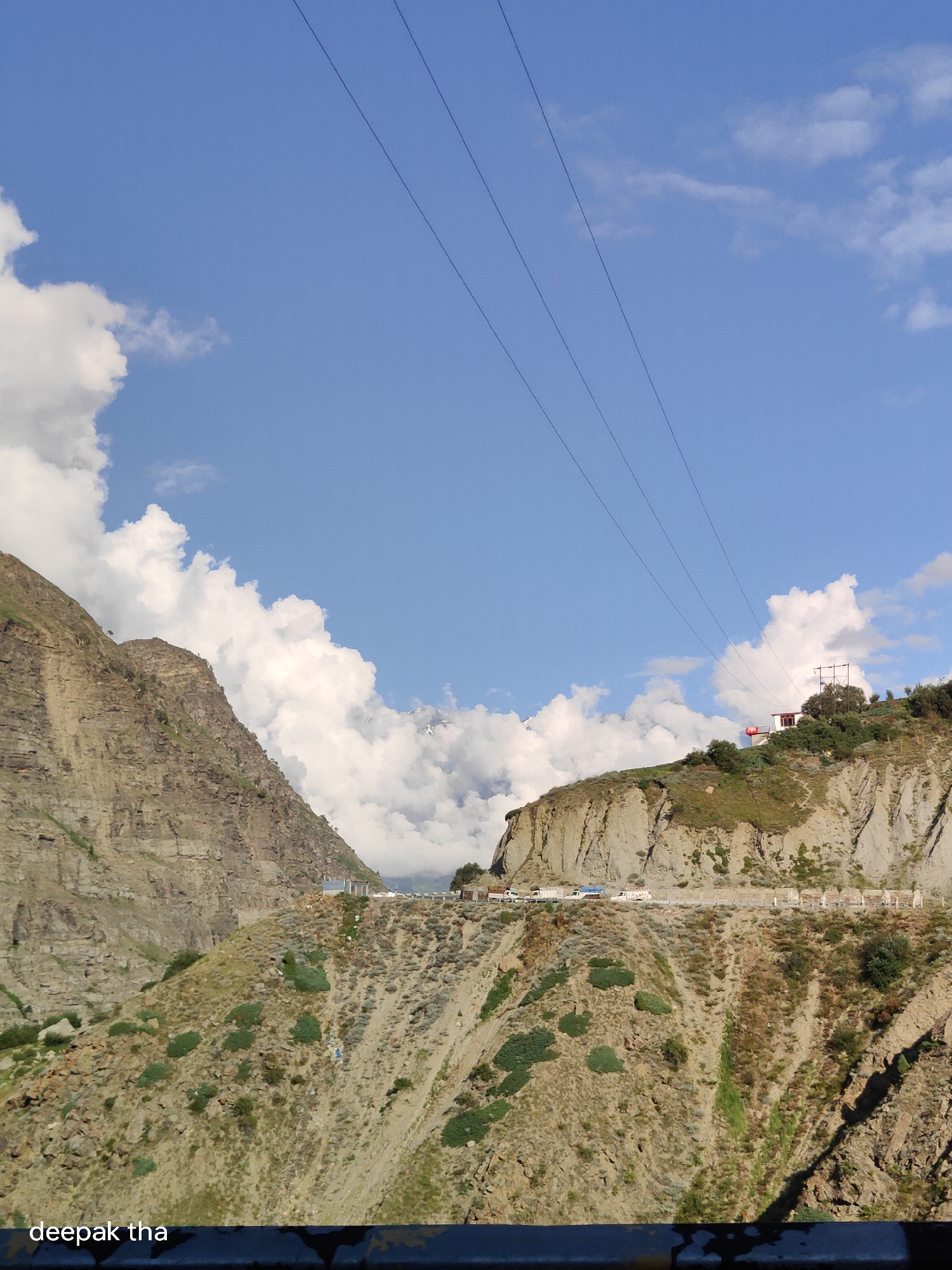 The image depicts a mountainous landscape under a bright blue sky dotted with fluffy white clouds. In the foreground, a steep, rocky mountainside covered with sparse vegetation slopes downwards. A road winds along the side of another, larger mountain in the background, with several vehicles, including trucks and a white van, visible on it. A building with a red roof is situated on the crest of the mountain. A series of power lines stretch diagonally across the sky from the upper left to the upper right of the frame. The lower portion of the image is partially obscured by a dark, horizontal railing. The text "deepak tha" is visible in the bottom left corner. The location is identified as Shuiling, India.