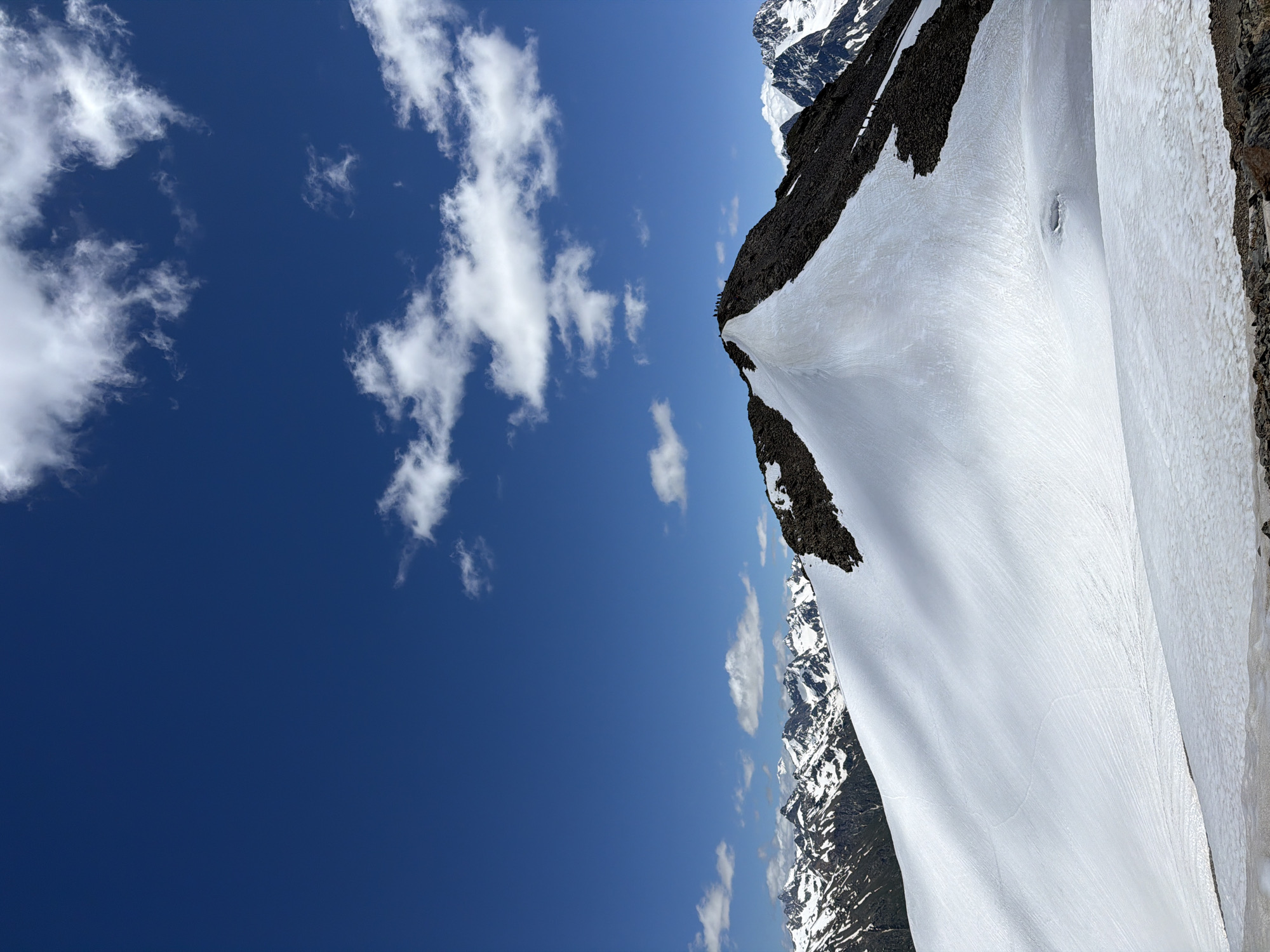 A high-angle, full shot captures a snow-covered mountain peak under a bright blue sky with scattered white clouds. The summit is adorned with dark, rocky terrain and a thin layer of snow, with a group of approximately ten people visible on the highest point. The foreground features a sweeping expanse of snow, marked by gentle slopes and subtle indentations, suggesting a recent thaw or glacial melt. In the distance, a chain of snow-capped mountains stretches across the horizon, their peaks sharp and jagged against the azure backdrop. The lighting suggests daytime, with shadows cast across the snowy surfaces, indicating a sunny environment. The scene is devoid of any urban structures or distinct city landmarks, implying a remote, natural mountainous setting.