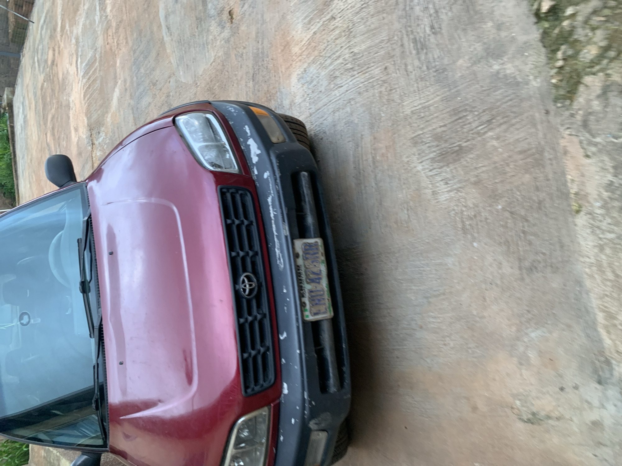 A red Toyota RAV4 is parked on a concrete surface outdoors. The car is facing the camera, and its front end, including the hood, grille, headlights, and bumper, is visible. A license plate with the characters "LBU-4ZSR" is attached to the front bumper. The concrete surface appears worn and uneven, with some patches of dirt and vegetation. The background is blurred, but appears to be a yard or open area with some greenery. The scene is overcast or in shadow, with soft, diffused lighting.