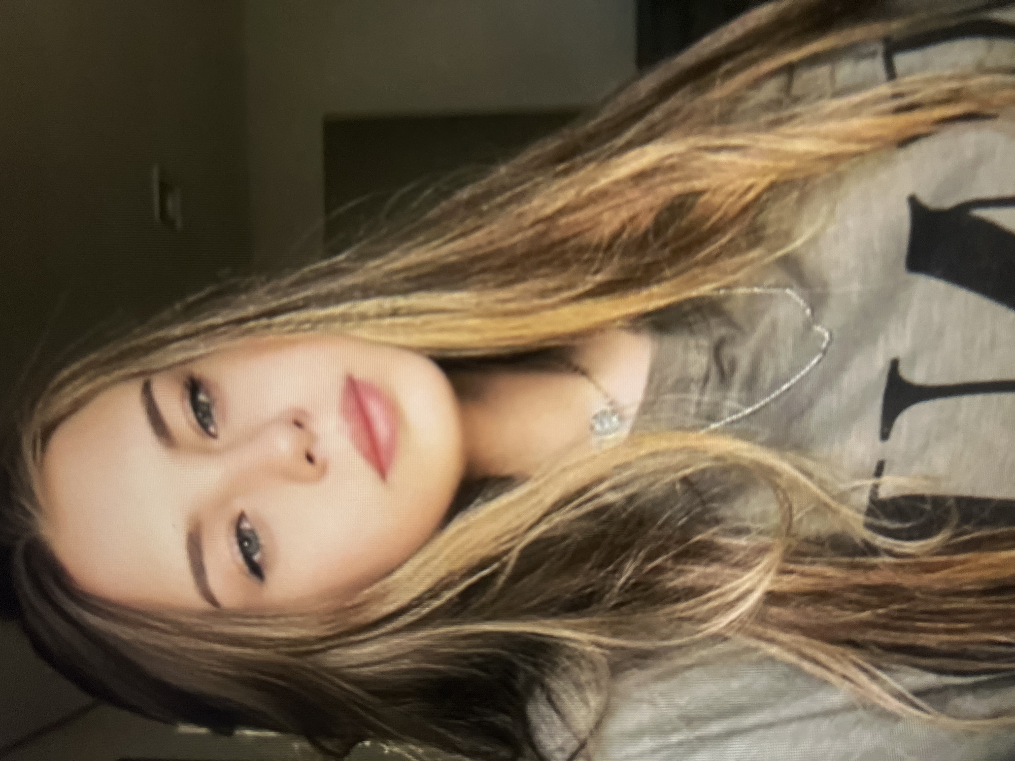 A close-up, waist-up, slightly blurry photograph shows a young woman with long, wavy, honey-blonde hair. She has light-colored eyes with winged eyeliner and pale pink lipstick. She is wearing a grey t-shirt with large black lettering, the full word of which is not visible, and a delicate silver chain necklace. The background is dark and out of focus, with some indeterminate shapes suggesting an indoor setting. There is no visible indication of the location in the city of Qarağandı city administration, Kazakhstan.