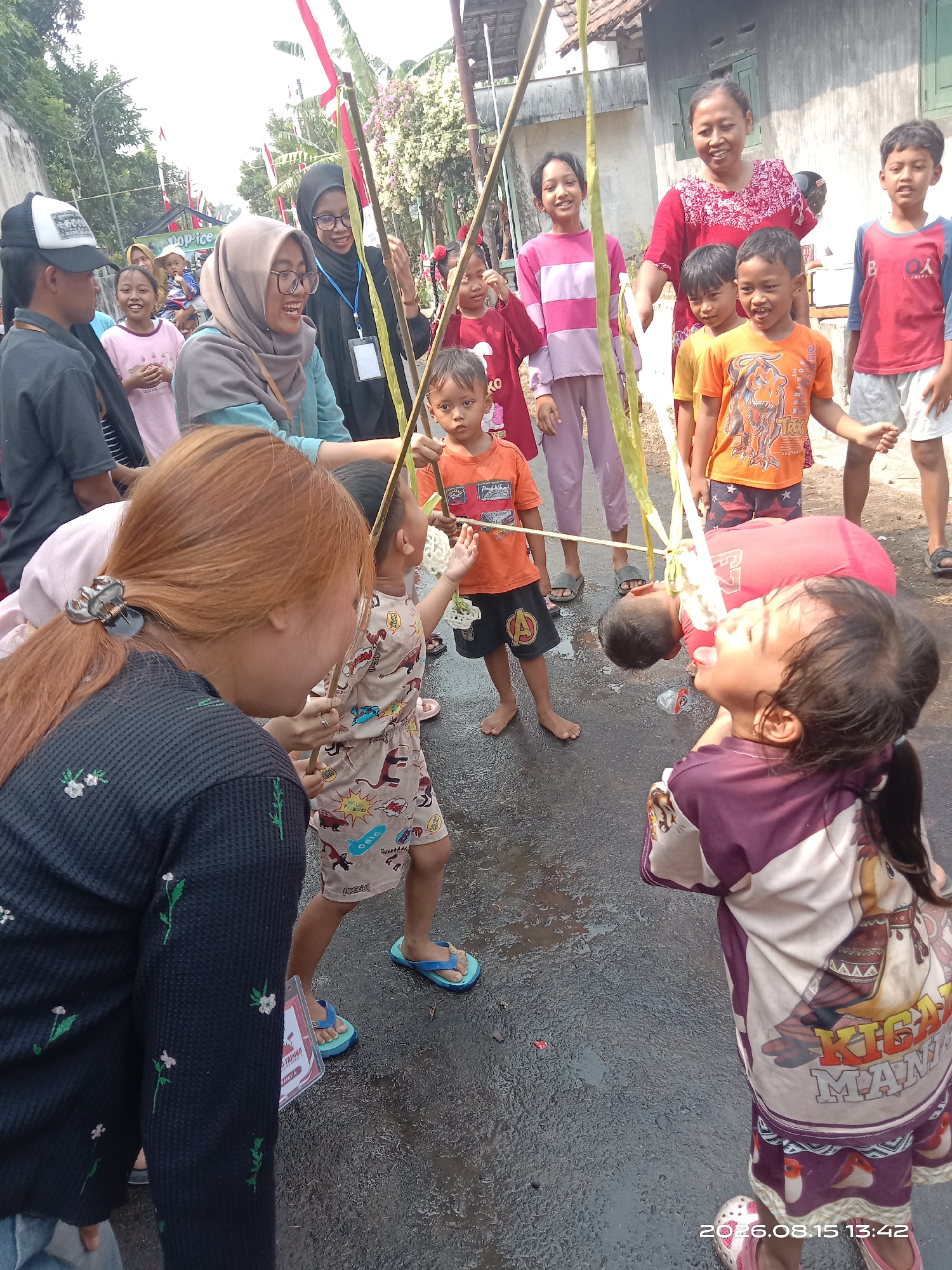 In Banyuarang, Indonesia, a group of children and adults are gathered on a wet, paved street, participating in an outdoor event. The scene appears festive, with Indonesian flags visible in the background. The central activity involves a game where two children, suspended by thin ropes tied to bamboo poles, are attempting to eat snacks. One child, wearing a red shirt, has their head tilted back, mouth open, reaching for a snack. A young girl in a purple and white patterned outfit stands in the foreground, looking towards the suspended child. Several other children and adults are observing the game. The environment suggests a community gathering, possibly a local celebration or festival, with a residential building visible in the background. The overall atmosphere is lively and engaged.