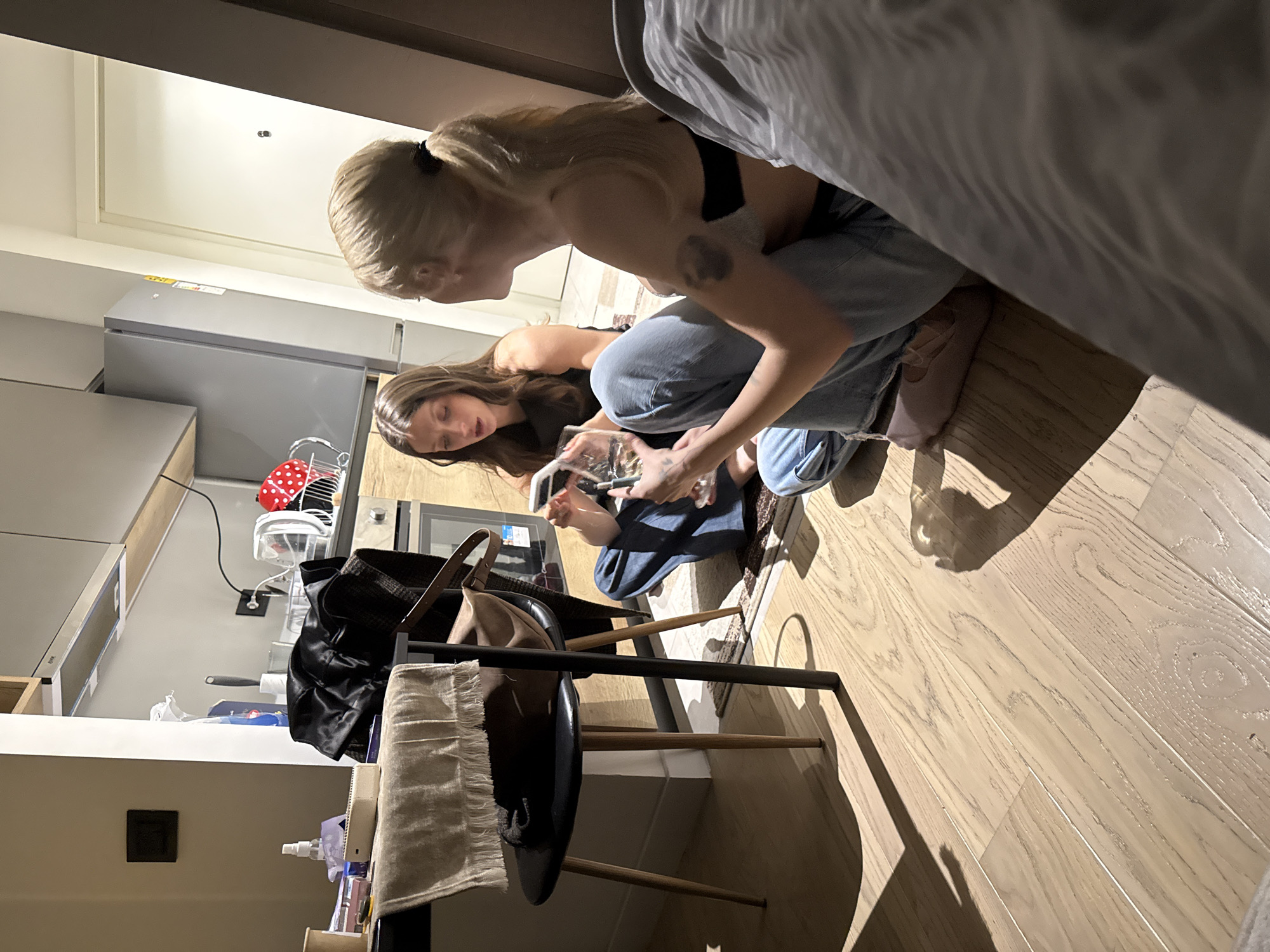 Two women are crouched on a light-colored wooden floor in what appears to be an apartment. One woman has blonde hair in a bun, a black top, blue jeans, and pink shoes. The other has long brown hair, a black top, and blue jeans. They are interacting with an object that could be a glass or a phone. A chair with a black seat and light-colored legs is visible, with a purse or bag and a towel hanging on it. The setting suggests an apartment kitchen or living area, based on the presence of kitchen appliances and furnishings. The location is unspecified beyond the general setting of an apartment, and the city of Belgrade, Serbia, cannot be confirmed from the image.