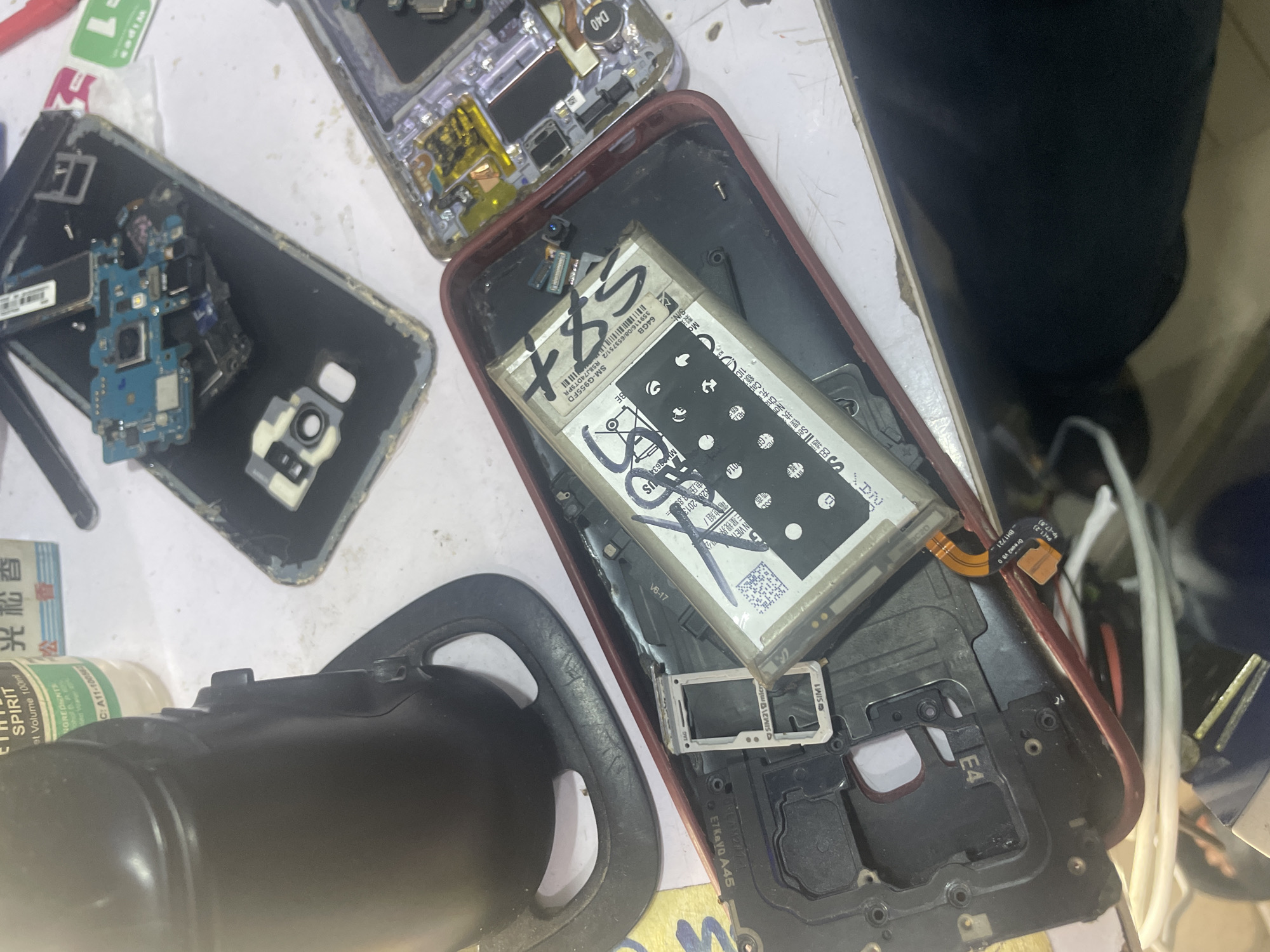 The image depicts a close-up, overhead view of electronic components, likely from disassembled mobile phones, laid out on a white surface. The scene appears to be a repair workshop or a workspace for electronics enthusiasts. Prominently featured are a black suction cup device, a disassembled smartphone frame containing a battery and SIM tray, and other scattered electronic parts including a motherboard. The environment suggests an indoor setting, possibly a table or workbench. There are no people visible in the image, and the overall impression is one of disarray or active work in progress on electronic devices. The specific location in Jalingo, Nigeria is not determinable from the visual information provided in the image.