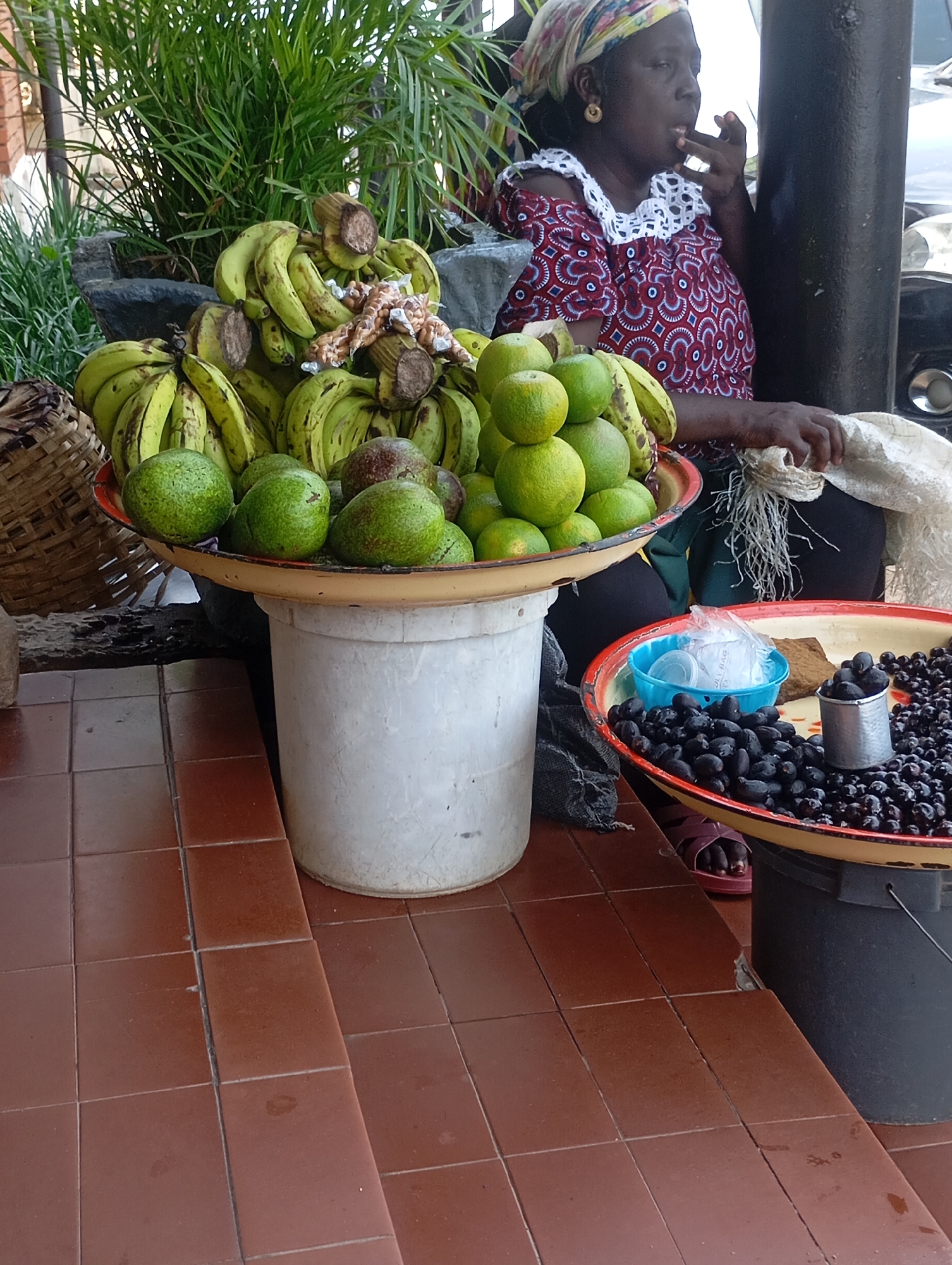 A dark-skinned woman, wearing a headscarf and a red and white patterned off-the-shoulder top with white ruffled trim around the neckline, is seated outdoors. She is holding a small object to her mouth, possibly eating. She is near a stall with various fruits for sale. In the foreground, a large white bucket supports a yellow-rimmed tray filled with green fruits, possibly avocados or limes, and bunches of green bananas. To the right, another yellow-rimmed tray contains a pile of small, dark, oval fruits, likely olives, and a small metal cup. A blue plastic container is also visible on this tray. A dark-colored bucket is placed below this tray. The ground is tiled with reddish-brown squares. The background features lush green foliage, a dark pole, and part of a vehicle. The overall scene depicts a market stall environment, consistent with a location in Jos, Nigeria.
