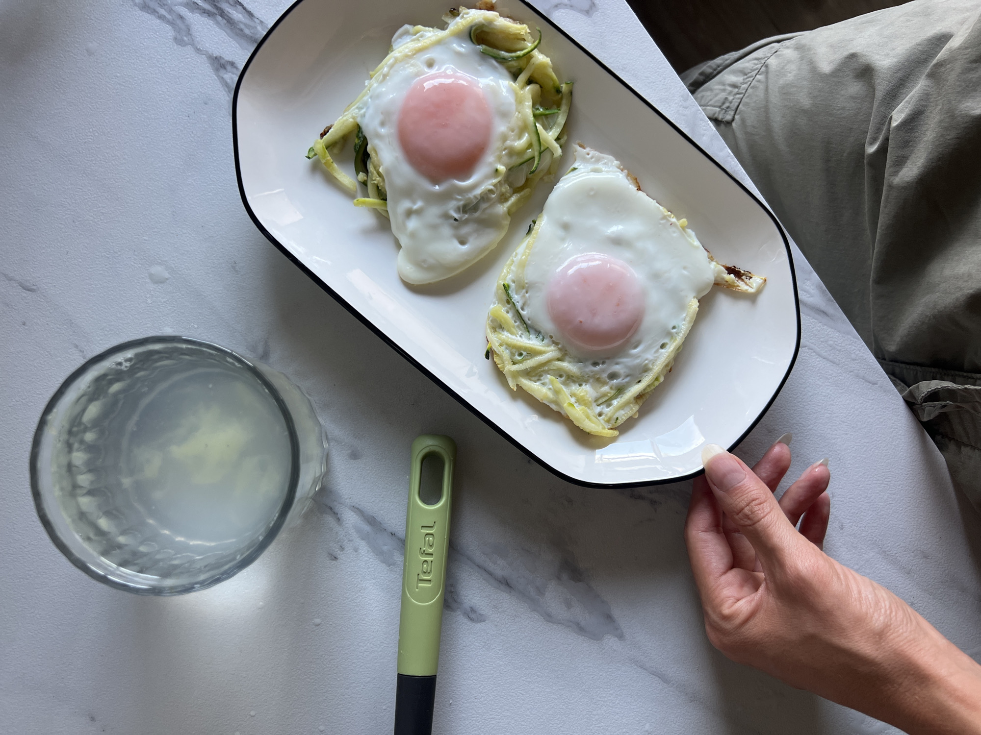 A overhead shot captures a close-up of a breakfast meal. Two fried eggs with pink yolks are served on a white oblong plate with a black rim. The eggs are placed atop a bed of shredded zucchini. To the left of the plate is a clear glass filled with a light-colored liquid. A light green utensil with the brand name "Tefal" is positioned near the glass. A person's hand is visible on the left side of the plate, their fingers delicately touching the edge. The setting appears to be a table with a white marble pattern.