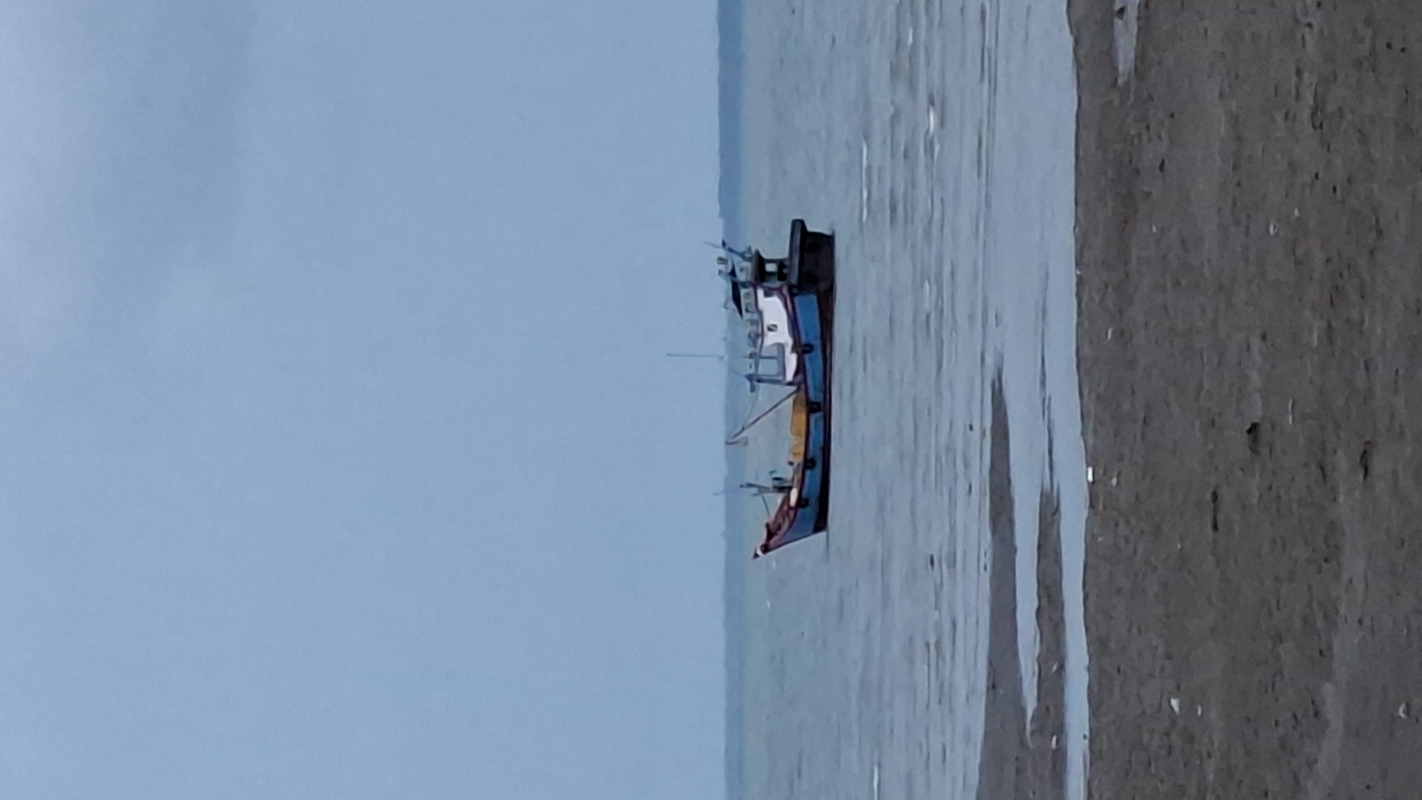 A fishing boat is moored in the water near the shore of Lhokseumawe, Indonesia. The boat is blue and white with red accents and has yellow netting visible on deck. The sky is overcast and a muted blue. The water is calm with small ripples. The foreground is a sandy, wet beach with some debris scattered on it. The scene appears to be a quiet day at sea, possibly during low tide.