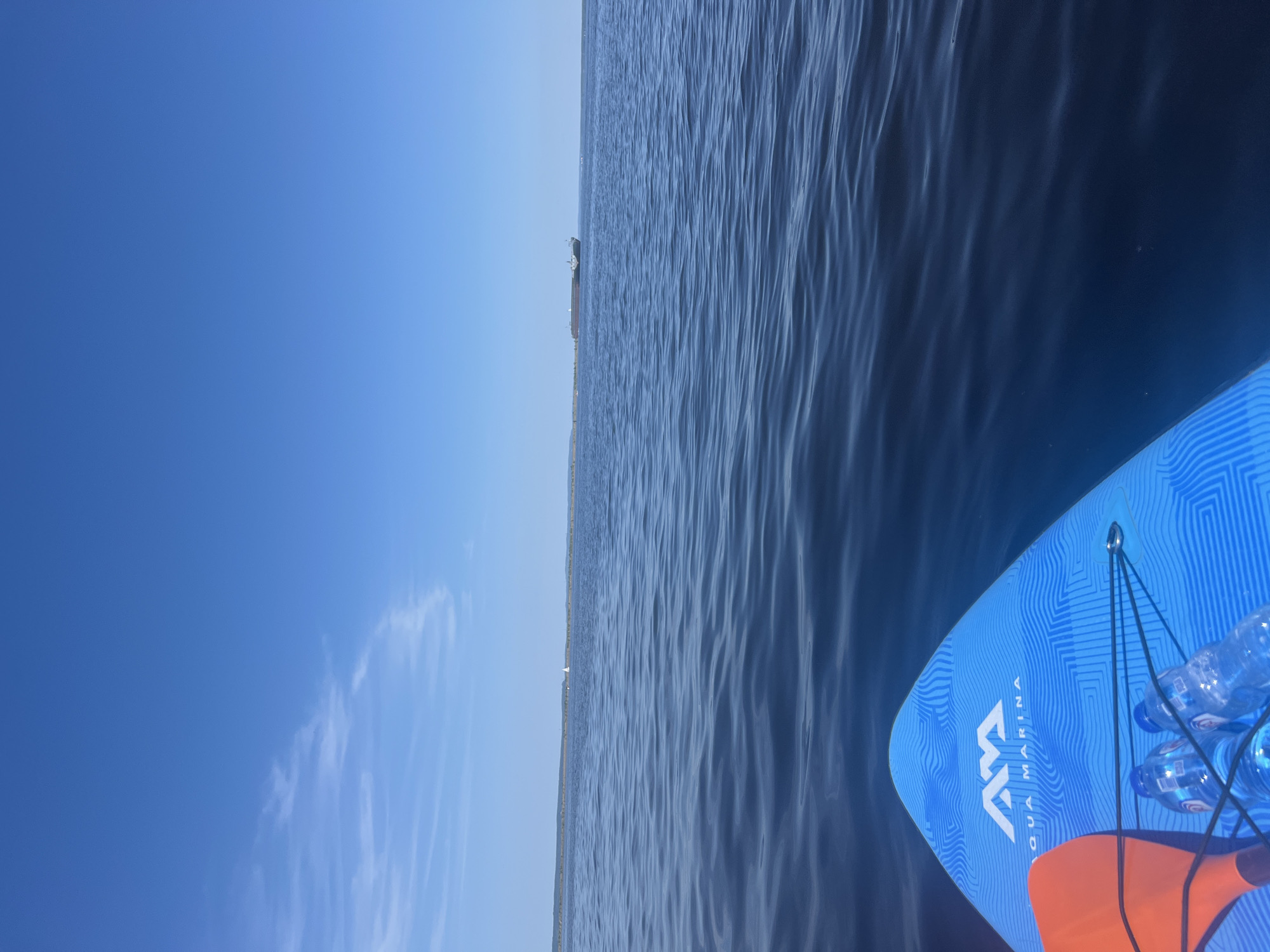The image depicts a first-person perspective from a paddleboard on a calm, dark blue body of water. The foreground shows the nose of a bright blue paddleboard with the "Aqua Marina" logo visible. An orange paddle and two plastic water bottles are secured to the board with black bungee cords. The middle ground features a long, thin strip of land or a breakwater stretching across the horizon, with some vegetation visible. A small white sailboat is near the left side of this breakwater, and a large, dark cargo ship is positioned further to the right, also on the horizon. The sky is a clear, bright blue with a few wispy white clouds on the left side. The water is smooth with gentle ripples. The location is the Kama River in Chaikovsky, Russia, a common spot for water activities. There are no people visible in the frame.
