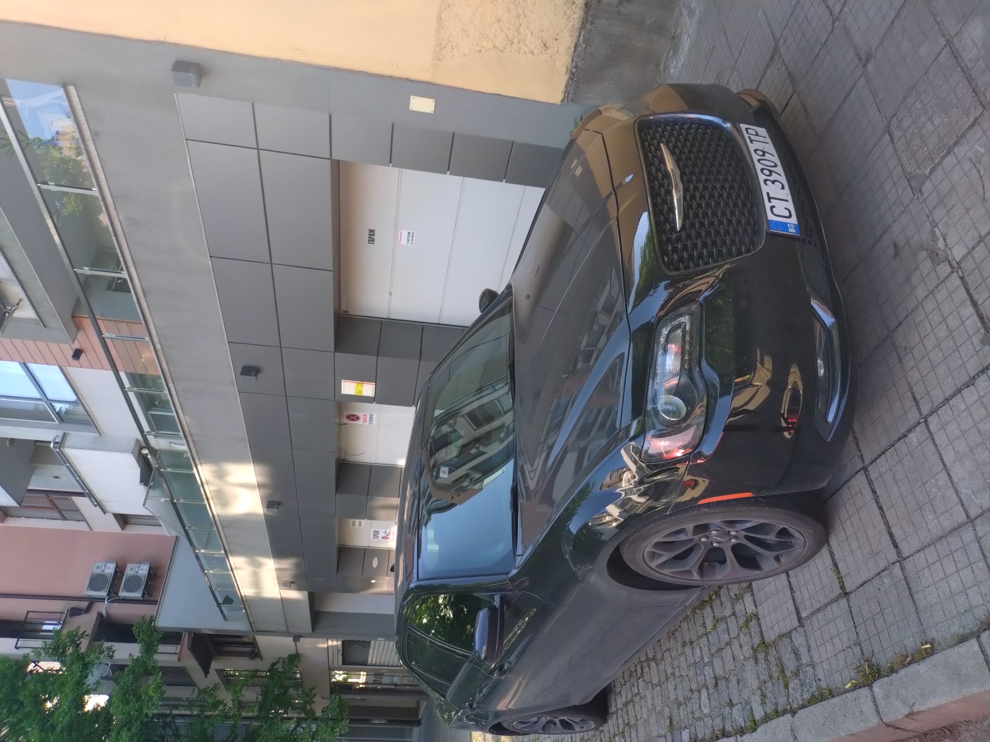 A black Chrysler 300 sedan, bearing Bulgarian license plate CT 3909 TP, indicating registration in Stara Zagora, is parked on a square-paver sidewalk. The vehicle is positioned in front of a multi-story building. The lower section of the building features a modern gray paneled facade with two white garage doors. Each garage door is labeled "ГАРАЖ" (GARAGE) in red Cyrillic text; one door also displays "НЕ ПАРКИРАЙ" (DO NOT PARK). Above the garage section, a balcony with a glass railing is visible. The upper stories of the building present a faded pinkish-brown facade, with balconies featuring metal railings and two external air conditioning units mounted on the wall. To the immediate right of the gray facade, an older, textured wall with a yellowish-beige hue is present. Green tree foliage is visible on the left side of the image, and the sidewalk shows minor grass growth between the concrete blocks. The scene is illuminated by natural daylight, casting shadows on portions of the building. No individuals are present.