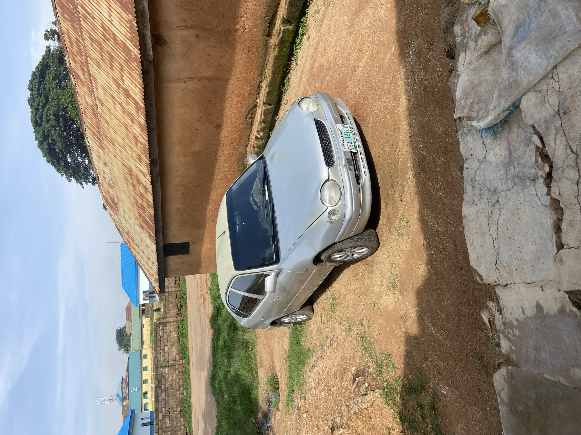 The image depicts a silver sedan parked on a dirt and gravel surface outside a building with an orange, textured wall and a rusted corrugated metal roof. The car's license plate, visible on the front bumper, reads "BSA-341CM PLATEAU." In the background, several other buildings are visible, some with blue roofs, and a large green tree stands behind the main building. The sky is partly cloudy. The scene appears to be in a residential or semi-urban area.