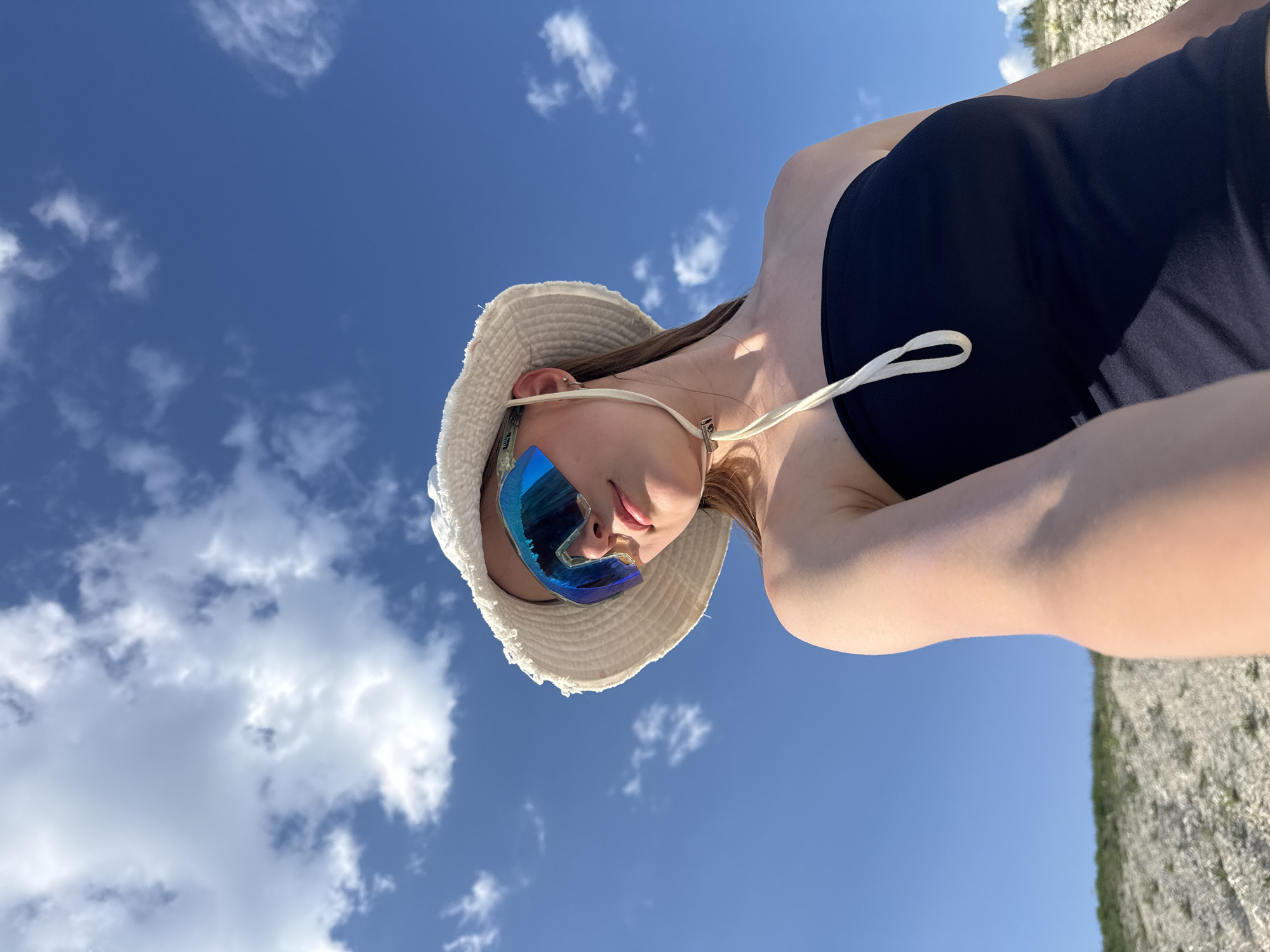 A low-angle shot depicts a person from the chest up, possibly in the city of Khvalynsk, Russia, given the request. The person is wearing a black top, a white bucket hat, and mirrored sunglasses. The background shows a blue sky with scattered white clouds. The edge of a light-colored, possibly stone or chalk, surface is visible in the lower right corner.