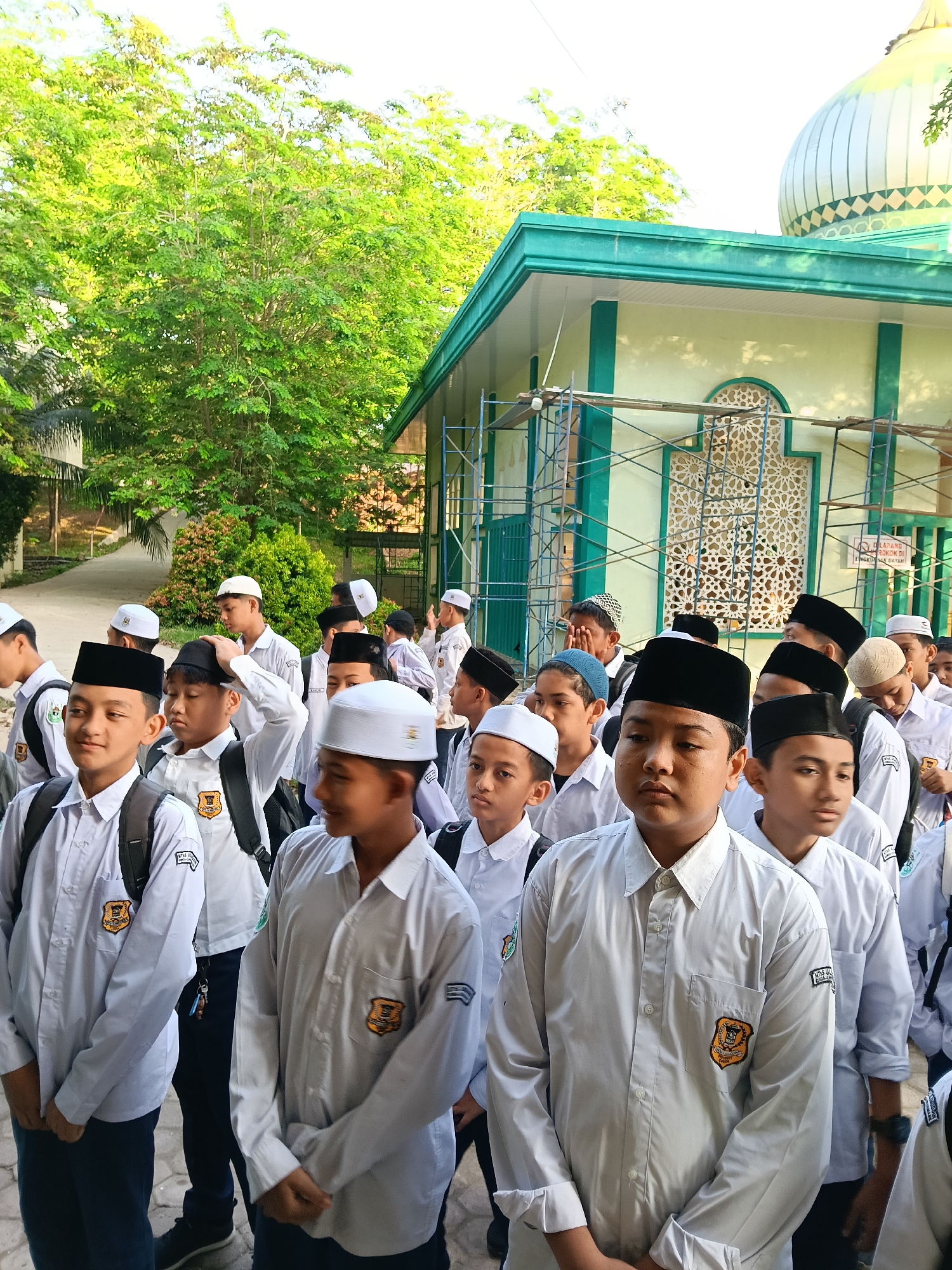 A group of male students, mostly wearing white shirts with school emblems, black caps, and some with white caps, are standing outside a building with a green and white dome, possibly a mosque, in Lhokseumawe, Indonesia. Some students have backpacks. Scaffolding is present on the side of the building, suggesting ongoing construction or renovation. The building has decorative latticework windows. Green trees are visible in the background.