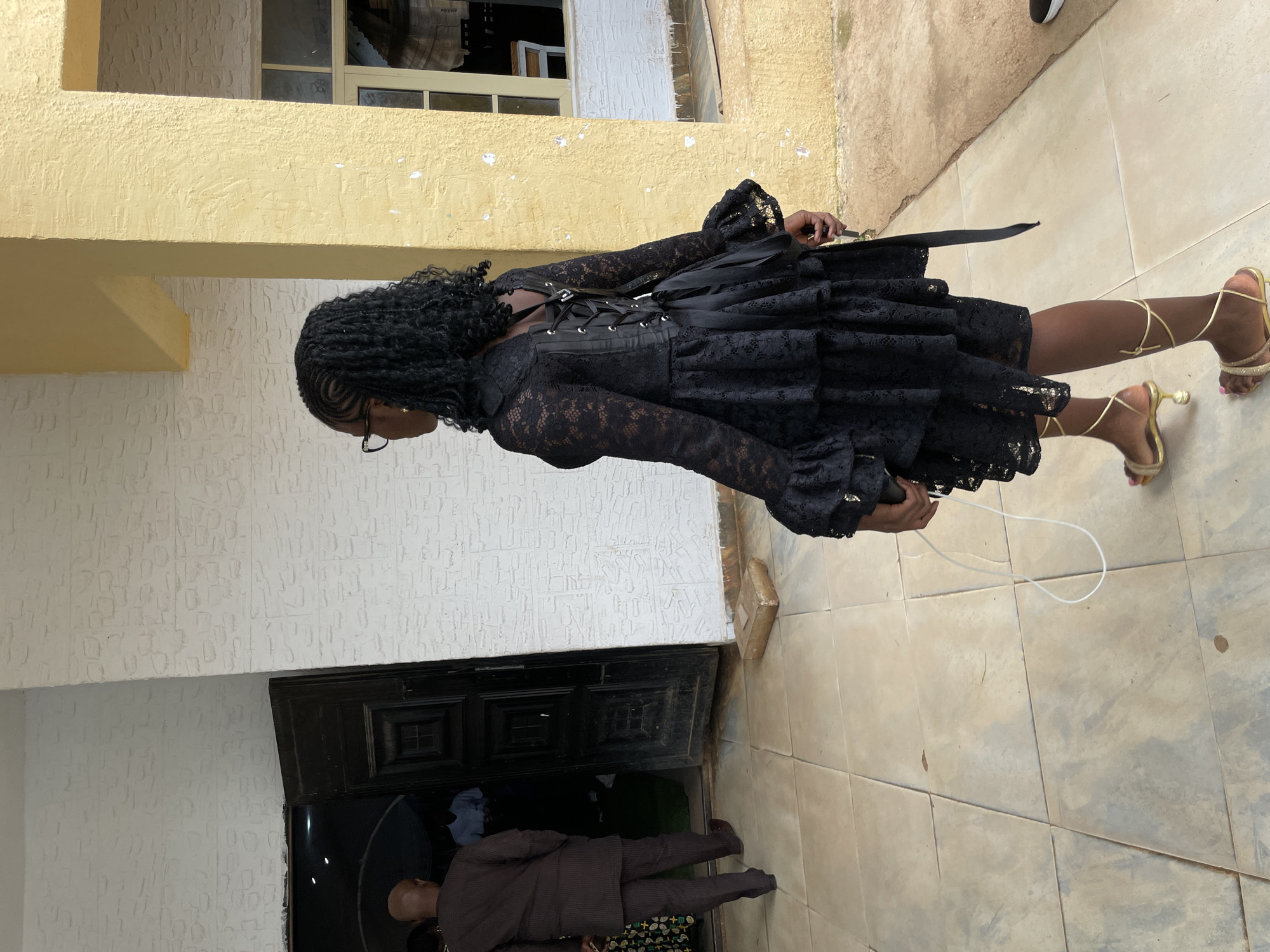 A young woman with dark, braided hair and glasses is walking away from the camera. She is wearing a black lace dress with a corset-like back and ruffled sleeves. Her sandals are gold and have straps that wrap around her ankles. To her left, a man in a brown pinstripe suit is walking towards an open doorway. Inside the doorway, a person in a light blue shirt is visible. The scene appears to be outdoors, with a textured white wall on the left and a yellow pillar on the right. The ground is paved with square tiles. This image was taken in Bar Gada, Nigeria.