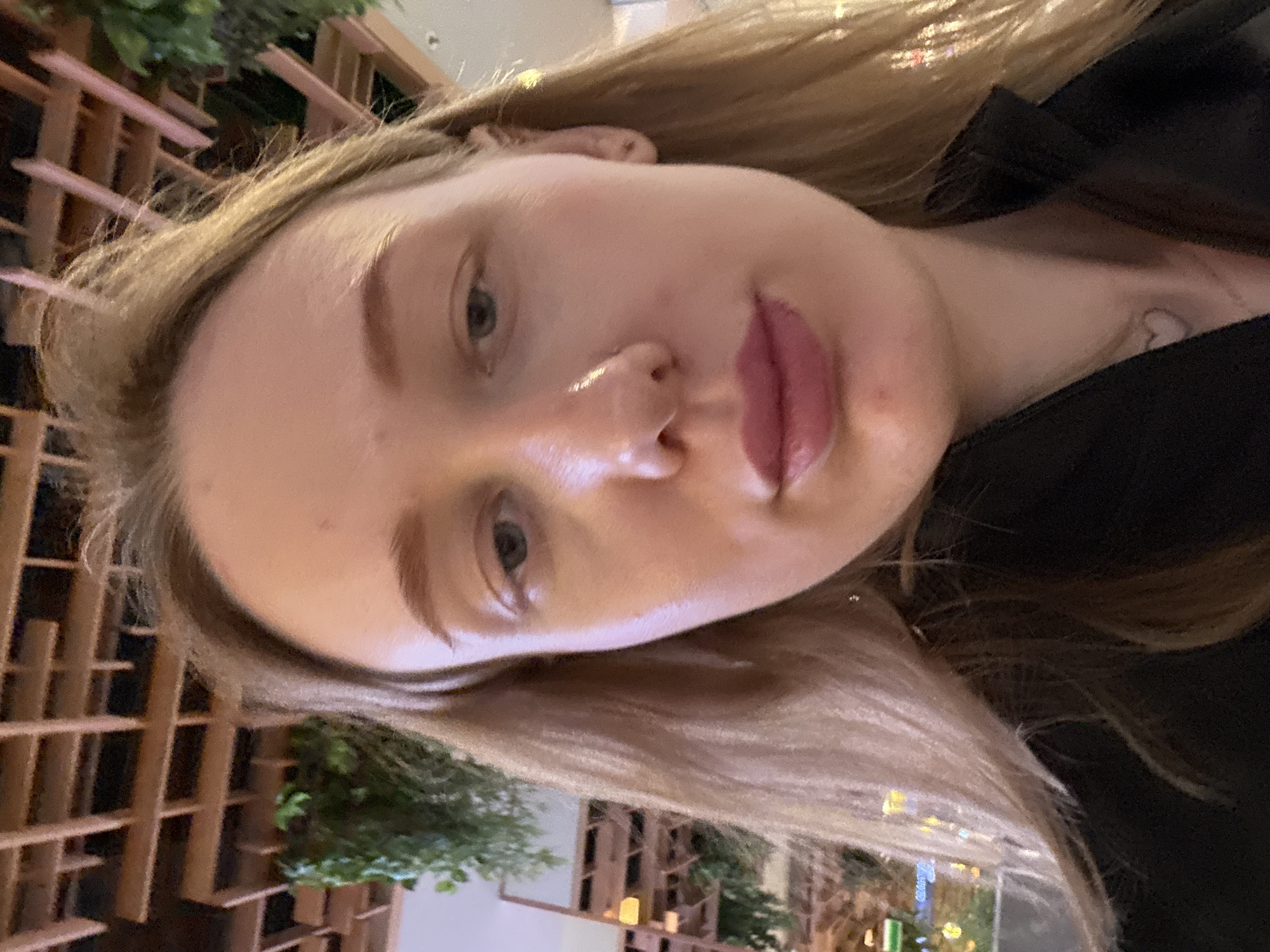 A close-up, rotated selfie of a fair-skinned woman with light brown hair. She is wearing a black top. The background appears to be an outdoor setting in Ol'ginskaya, Russia, suggested by the presence of a lattice-style fence with greenery and distant lights.