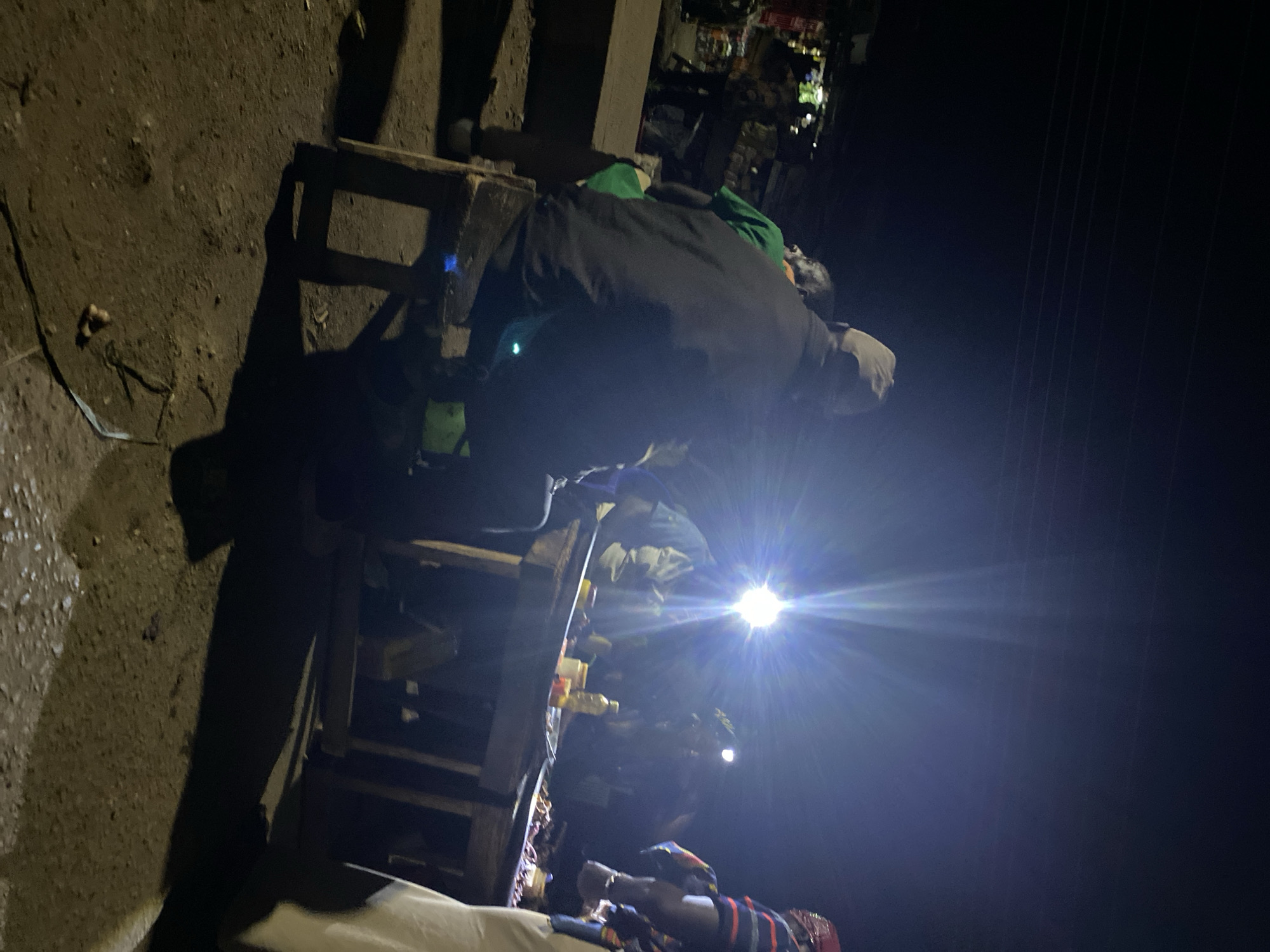 The scene shows an outdoor night market in Bukuru, Nigeria, illuminated by bright lights. A person wearing a dark jacket, possibly leaning over a wooden market stall, is visible. The stall displays various goods, including what appear to be bottles. Another individual lies on a surface, possibly covered by fabric with striped patterns. The ground appears to be earthen. The background is obscured by darkness.