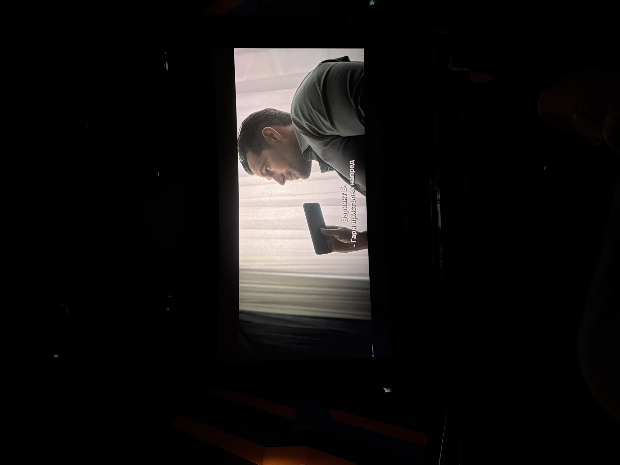The image depicts a scene inside a dark cinema hall, focusing on a large screen displaying content. On the screen, a man with short dark hair and a beard is shown in profile, looking down intently at a black smartphone he holds. He appears to be wearing a dark or olive-colored collared shirt or jacket. The man's face and the phone are brightly illuminated by the screen, contrasting with the overall dark environment.
White text in Cyrillic is visible on the screen, acting as subtitles. The text reads: "Вариант Б." (Variant B.) and below it, "- Гари пристъпва напред" (- Gary steps forward).
The surrounding cinema hall is predominantly dark, but some details are discernible. In the foreground and lower section of the image, the outlines of tiered cinema seating, possibly with orange or brown accents, can be vaguely seen. In the darker areas around the screen, particularly in the upper right, a green "EXIT" sign is faintly visible, confirming the setting as a cinema or similar venue. The overall atmosphere suggests an evening or night viewing session. The primary activity shown is the viewing of a film or television show on a large screen, with a character engaging with a smartphone within the on-screen narrative.