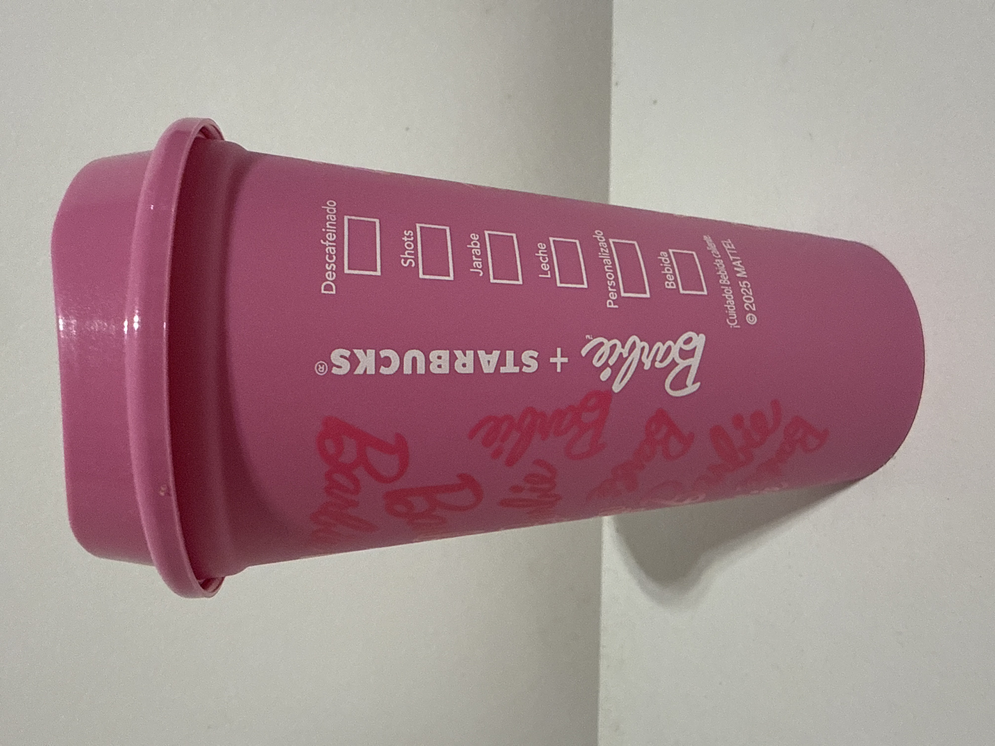 A pink plastic reusable Starbucks cup is shown resting on a white surface. The cup has a pink lid with a grooved edge. The cup's body has white printed text. The text includes the Starbucks logo, the word "Barbie" repeated in a pattern, and a section with checkboxes labeled "Descafeinado," "Shots," "Jarabe," "Leche," "Personalizado," and "Bebida." It also says "¡Cuidado! Bebida caliente ©2025 MATTEL." The setting appears to be indoors due to the presence of a plain white surface. The city of Buenos Aires, Argentina, is not visible or implied in the image.