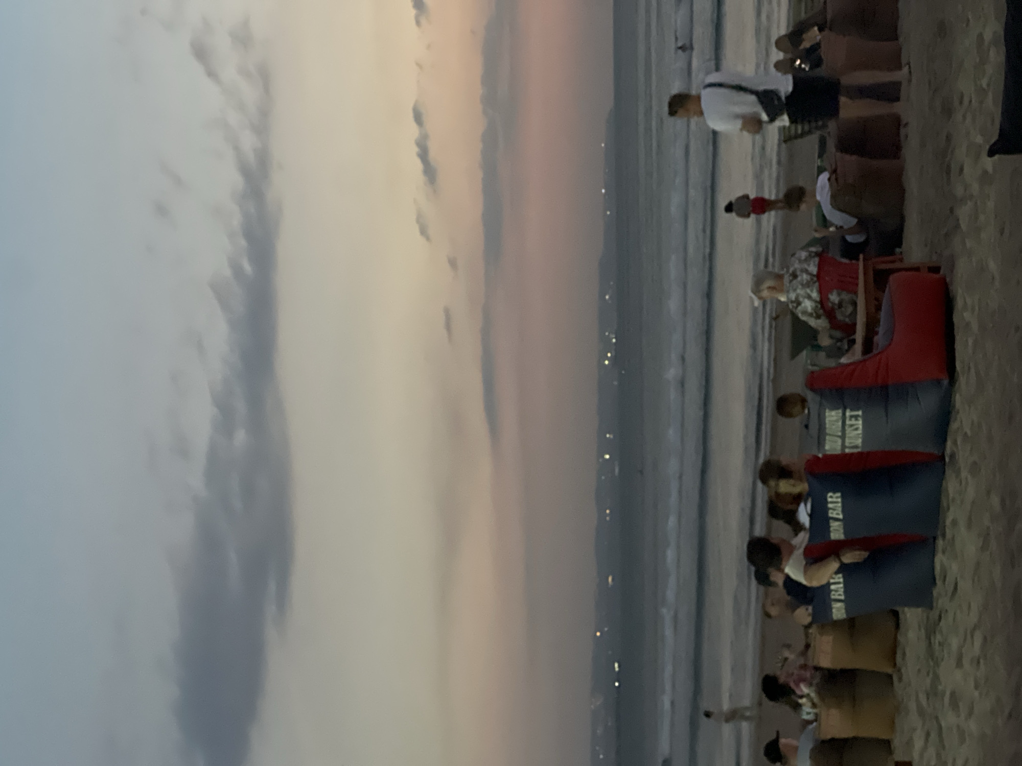 The image captures a twilight scene on Kuta Beach in Indonesia. Along the sandy shoreline, numerous people are scattered about, some seated on beach chairs. A few individuals are standing, and others appear to be walking near the water's edge. The sky displays a gradient of colors, with the sun setting. Notably, beach chairs with the words "IRON BAR COLD DRINK SUNSET" are evident.