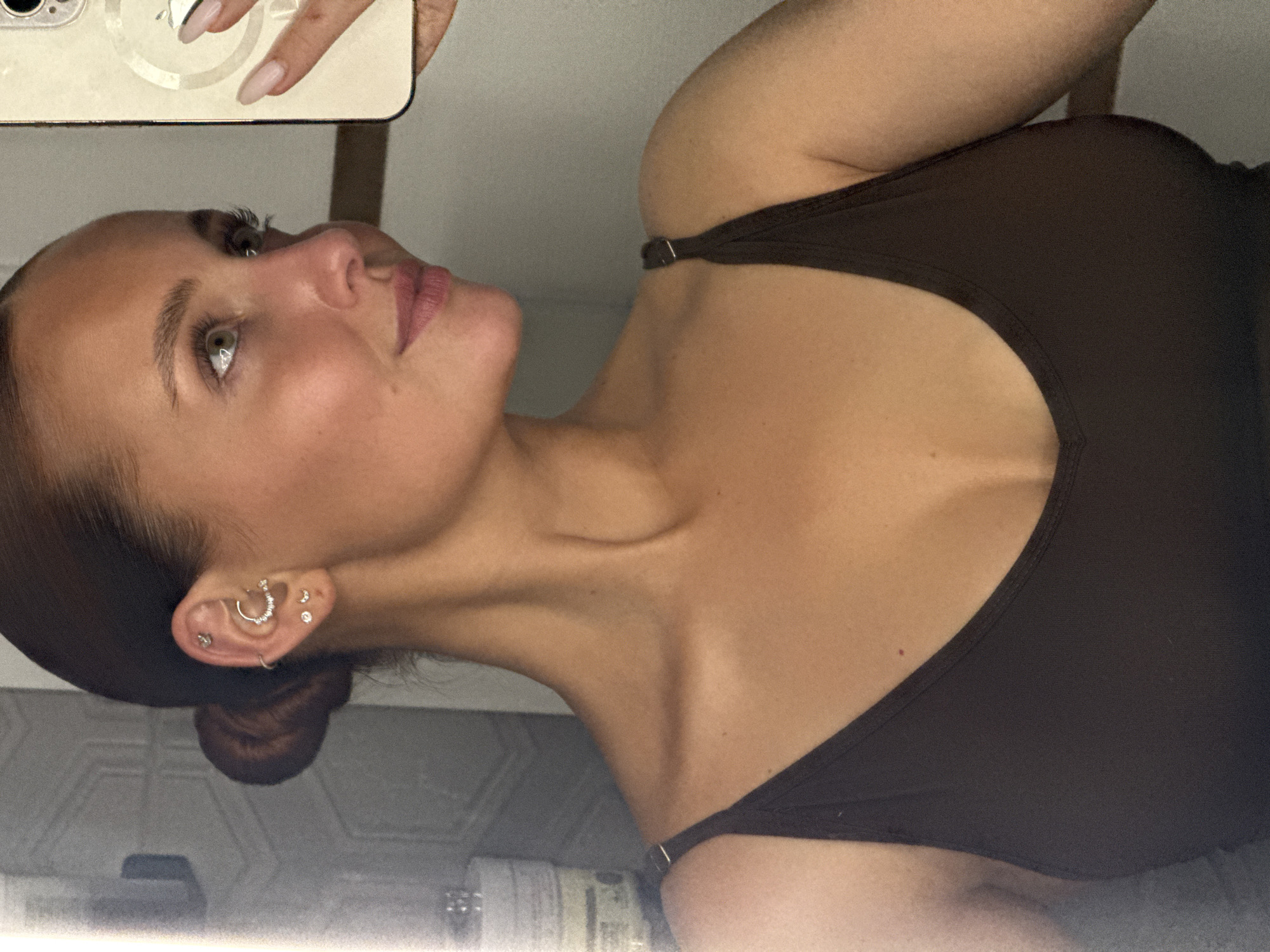 A woman is taking a selfie in what appears to be a bathroom. She has fair skin, light eyes, and dark hair pulled back into a bun. She is wearing a dark brown tank top. Her left ear has multiple piercings, including a small hoop and studs. She is holding a phone with a reflective surface in her left hand, which partially obscures the view. The background includes a tiled surface, possibly a wall, and some toiletries. It is not possible to determine the location as Chaikovsky, Russia, from the image alone.