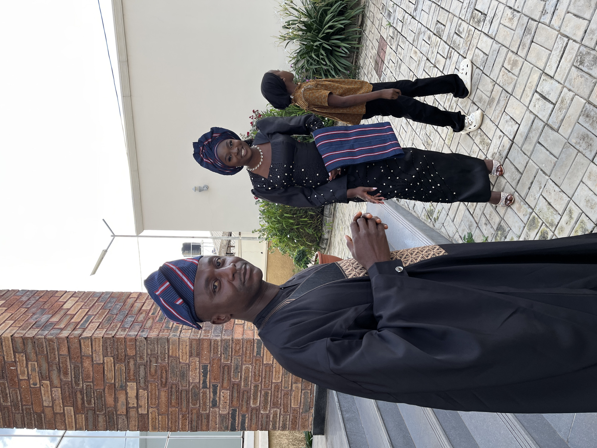 In Gura Top, Nigeria, a man dressed in traditional black attire with a patterned cap stands in the foreground, facing the camera with a slight smile. He is positioned on a paved walkway next to a brick building with large windows. Behind him, a woman in a black, beaded dress and a matching headwrap is seen from the waist down, holding a striped cloth. To her right, a child in a brown shirt and black pants walks away from the camera, also on the paved walkway. The environment appears to be an outdoor courtyard or patio with some greenery and a plain wall in the background.