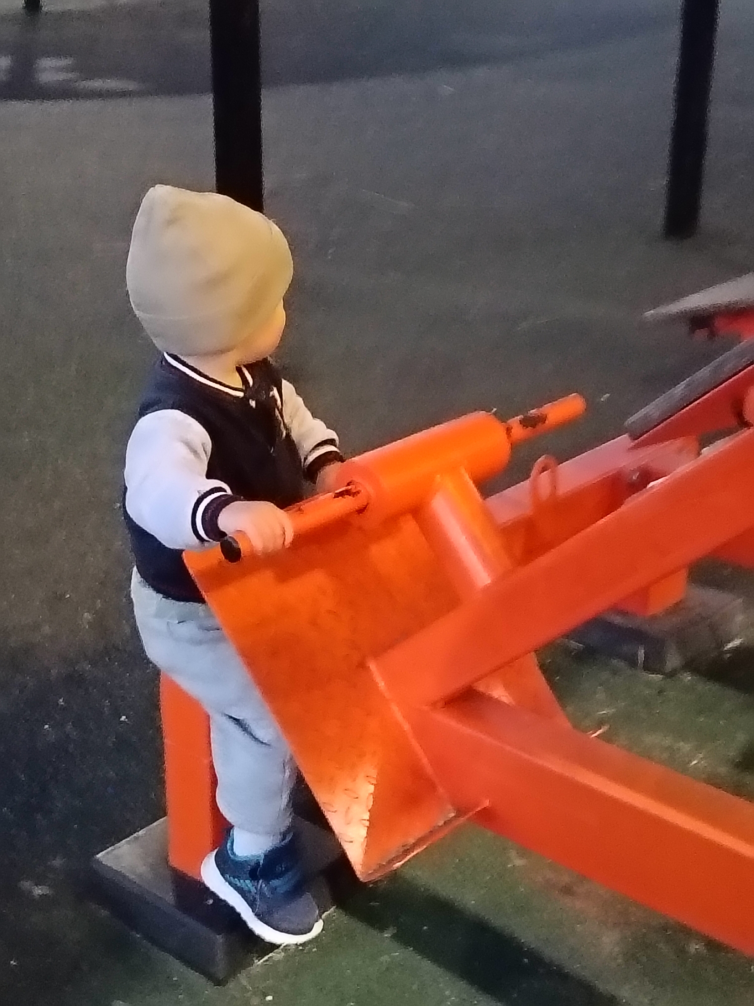 In Sudogda, Russia, a toddler wearing a light-colored beanie, a black and white jacket, grey pants, and dark sneakers stands on a black base. The child is positioned to the left of a bright orange playground apparatus with metal bars and cylindrical handles. The toddler's right hand is gripping one of the orange handles, and they appear to be looking towards the right. The ground is covered in dark, textured material, possibly rubber mulch or asphalt. A dark vertical pole is visible in the background to the left. The scene is outdoors, and the lighting suggests it is daytime or early evening.