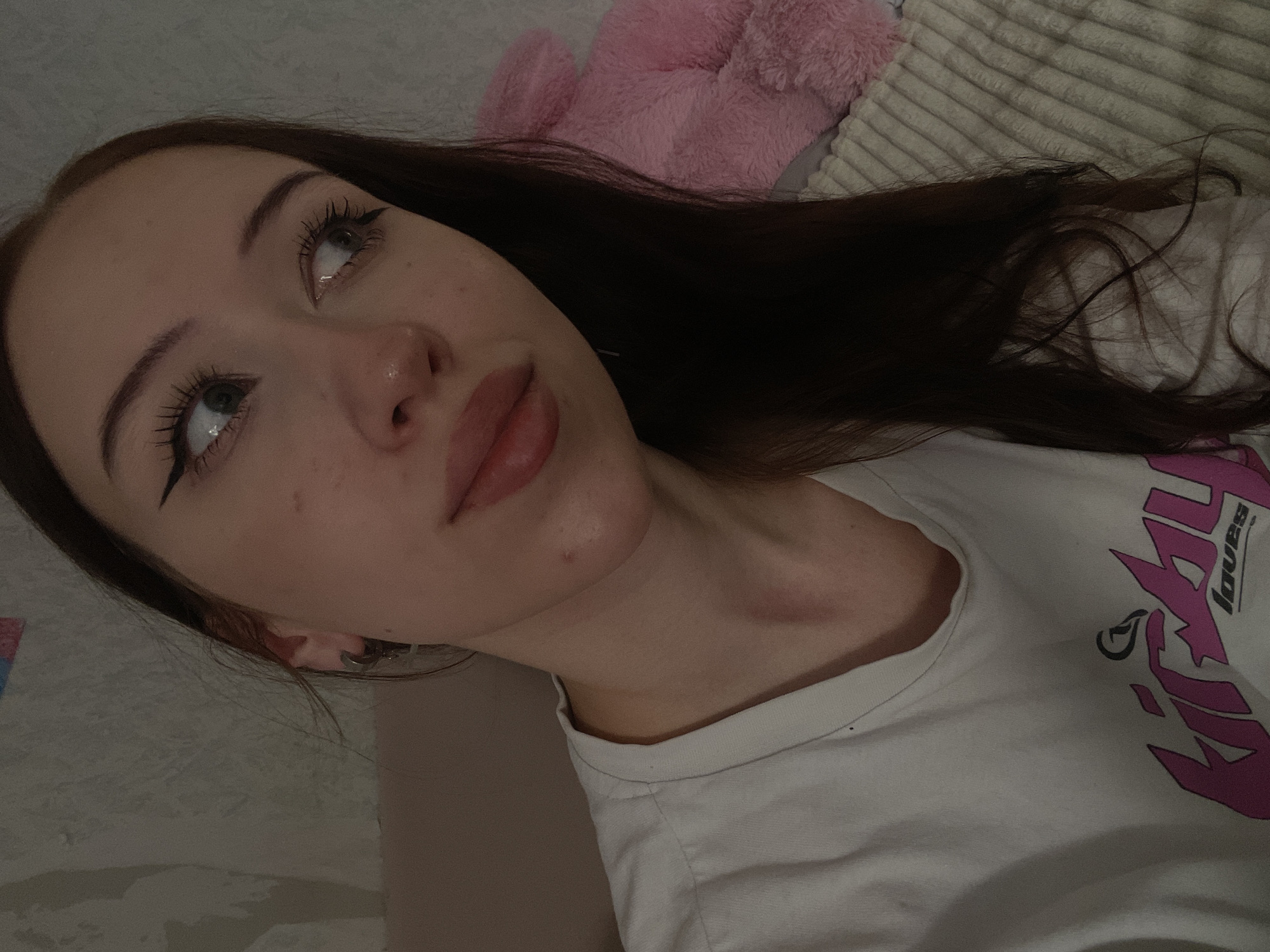 The image shows a close-up, head-and-shoulders view of a young woman with long, dark brown hair. She is wearing a white t-shirt with a pink graphic design. Her makeup includes winged eyeliner and mascara. She has light-colored eyes and fair skin with freckles. She is looking to her right, away from the camera. A pink stuffed animal is partially visible behind her. The background consists of a textured wall and a ribbed blanket. The scene is indoors, likely a bedroom. There is no visible indication of the city of Votkinsk, Russia.