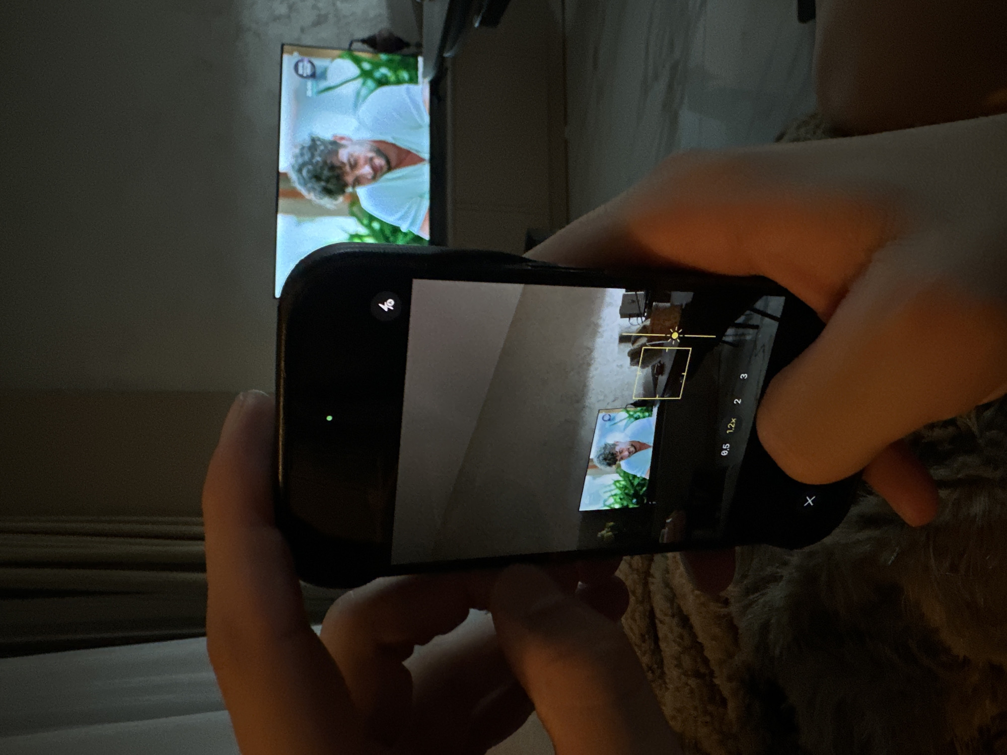 The scene depicts an indoor environment in Sofia, Bulgaria, captured from a first-person perspective. A person's light-skinned hands are holding a black smartphone, which is actively displaying its camera interface. The phone's screen presents a live preview of the room, prominently featuring a large, wall-mounted or placed flat-screen television. The television, visible both directly in the background and within the phone's camera preview, displays a male subject with curly dark hair and a beard, wearing a light-colored collared shirt, smiling. Green foliage is visible behind the male subject on the television screen. The smartphone's camera interface shows a disabled flash icon, a green indicator dot in the upper right (denoting active camera/microphone), zoom level options (0.5x, 1x, 2x, 3x), and a focus/light meter box in the lower right of the preview. The background consists of a light-colored wall, partially visible curtains (one light, one darker), and a dark, low-profile piece of furniture beneath the television. The surface beneath the hands and phone is covered with a light brown, textured, fluffy material. The room is dimly lit.