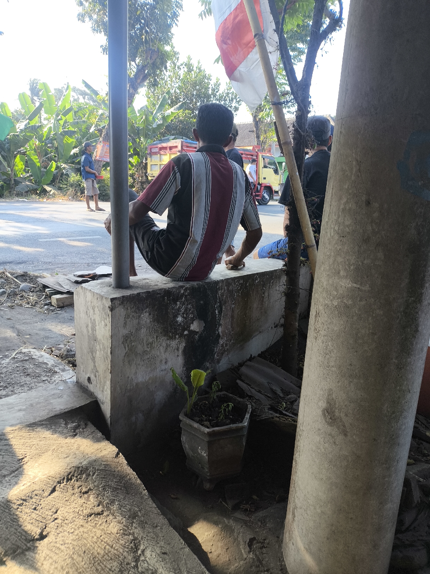 The image depicts an outdoor scene in Labruk Kidul, Indonesia, under bright daylight conditions. In the foreground, a person is seated on a concrete block, facing away from the camera, wearing a striped shirt and dark shorts. To the left, a second person stands near a road, holding a red object, dressed in a blue shirt and light-colored shorts. Behind them, a yellow truck is visible on the paved road, which is bordered by lush green banana trees and other foliage. A thin bamboo pole with a red and white flag is positioned vertically, partially obscuring the view of a tree with a thick, textured trunk to the right. The ground around the concrete block is unpaved and littered with debris.