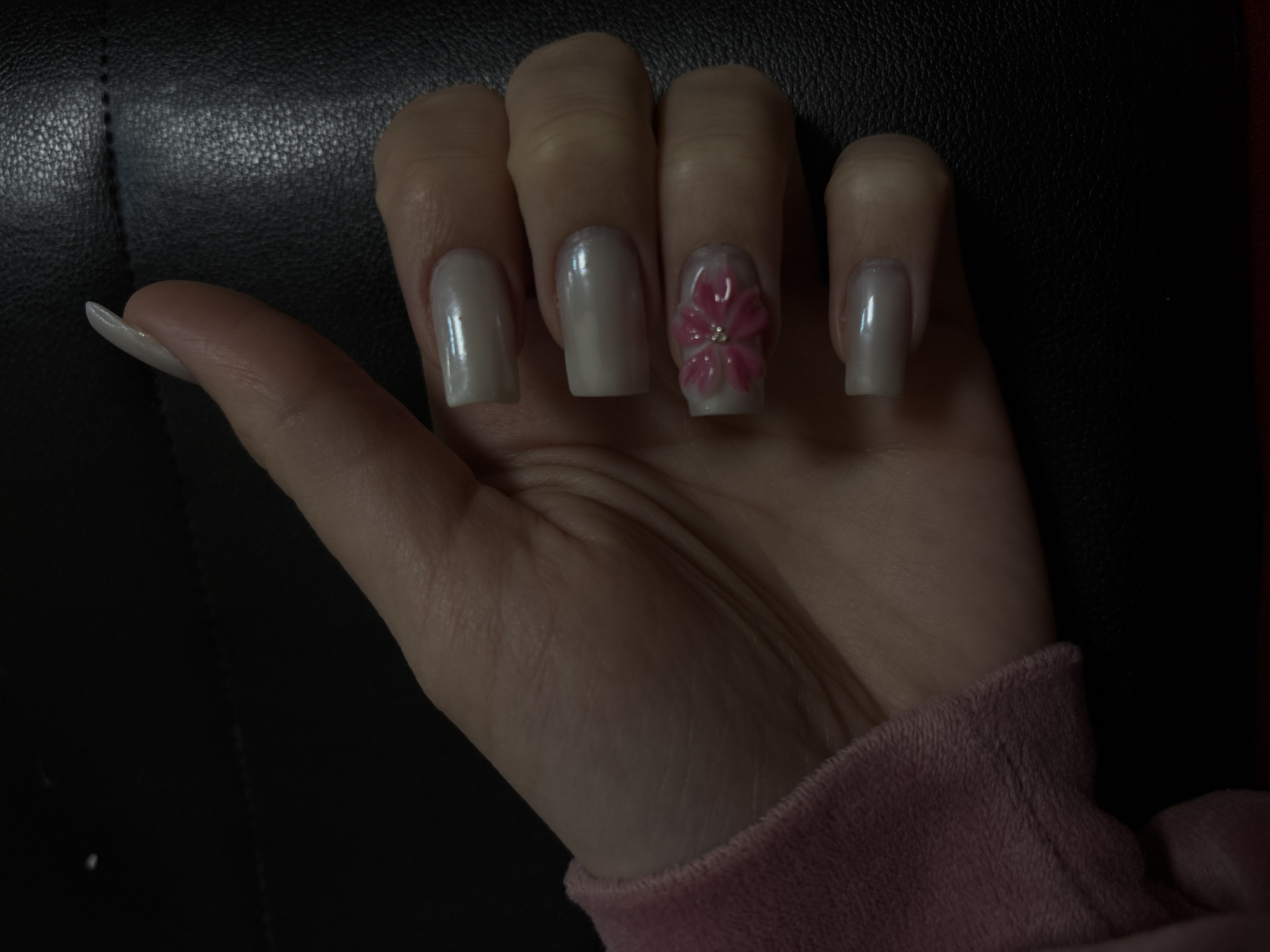 A medium shot depicts a light-skinned person's hand against a dark, textured surface. The hand has long, manicured nails. The nails are a pale, pearlescent color. The nail on the middle finger has a raised, pink flower design with a small, silver accent in the center. The hand is positioned at an angle, with the thumb pointing slightly to the left. The person is wearing a pink, plush garment at the bottom right corner. There are no other people or identifiable objects in the image. The location is not discernible from the image.