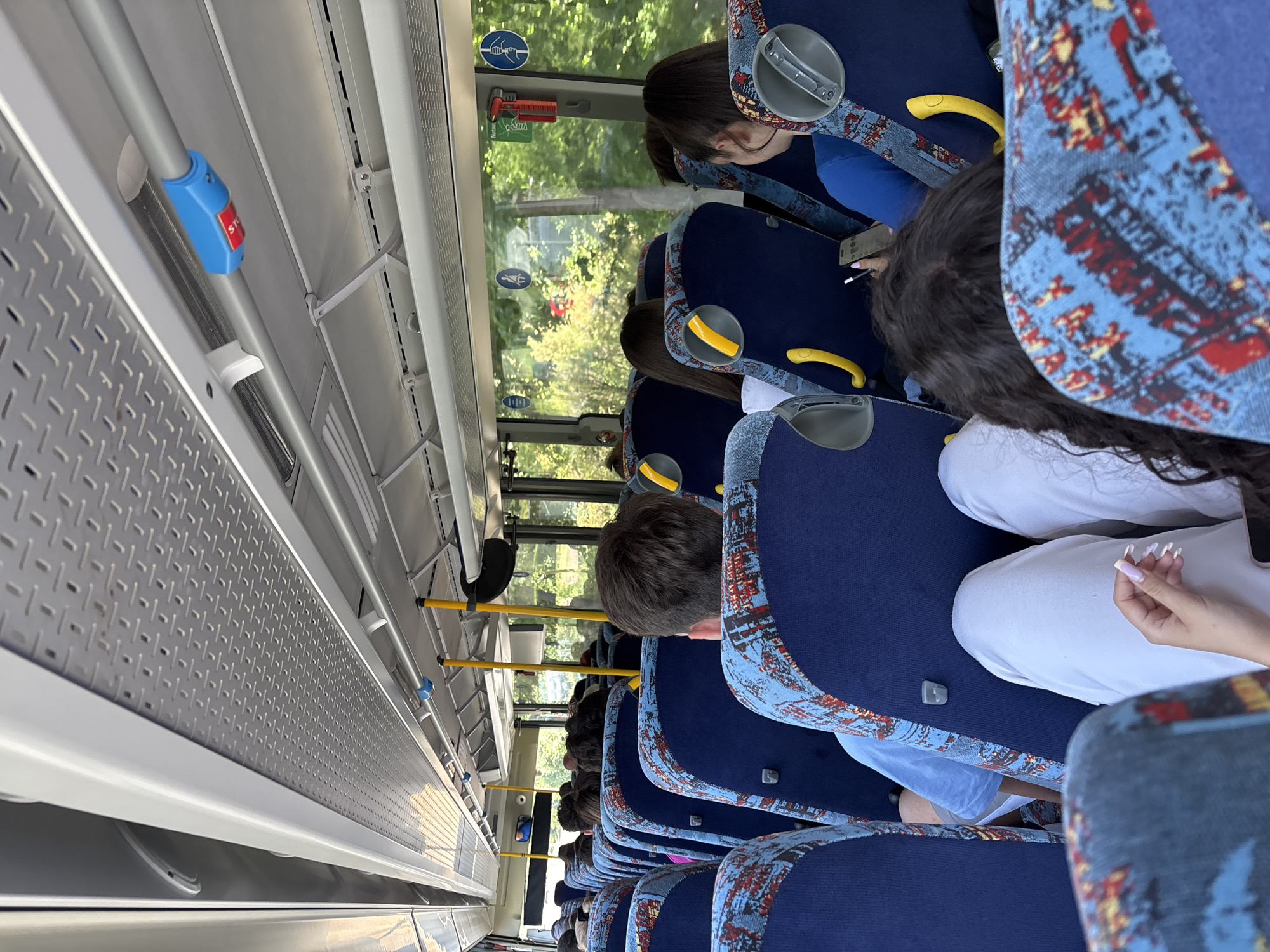 The image shows the interior of a public transport bus, viewed from a slightly elevated position looking forward. Several passengers are seated in rows, primarily visible from behind. The bus seats are blue with distinct patterned fabric accents in light blue, red, and yellow.
Along the left side of the bus, a long, textured metal panel runs above the window line. Below this, silver handrails extend, one of which has a blue casing with a prominent red "STOP" button. Yellow handrails are also visible further into the bus, interspersed among the seating. White support structures are mounted to the ceiling and side panels.
Through the windows on the right, abundant green foliage and trees are visible, indicating bright, sunny daytime conditions outside. Near the top of one window, two clear pictograms are displayed: one showing an emergency hammer breaking glass, and another depicting a person running towards an exit, presumably part of emergency instructions. Below these, a partially visible, vertically oriented word, likely "Notfall" or "Notaus," can be deciphered.
Passengers are generally obscured, but one individual near the front appears to be looking down, possibly at a phone. In the foreground, the hand of a passenger with long, light-colored manicured nails is visible, resting on their lap. The scene depicts a routine journey on public transportation, consistent with an urban or suburban setting in Burgas, Bulgaria.