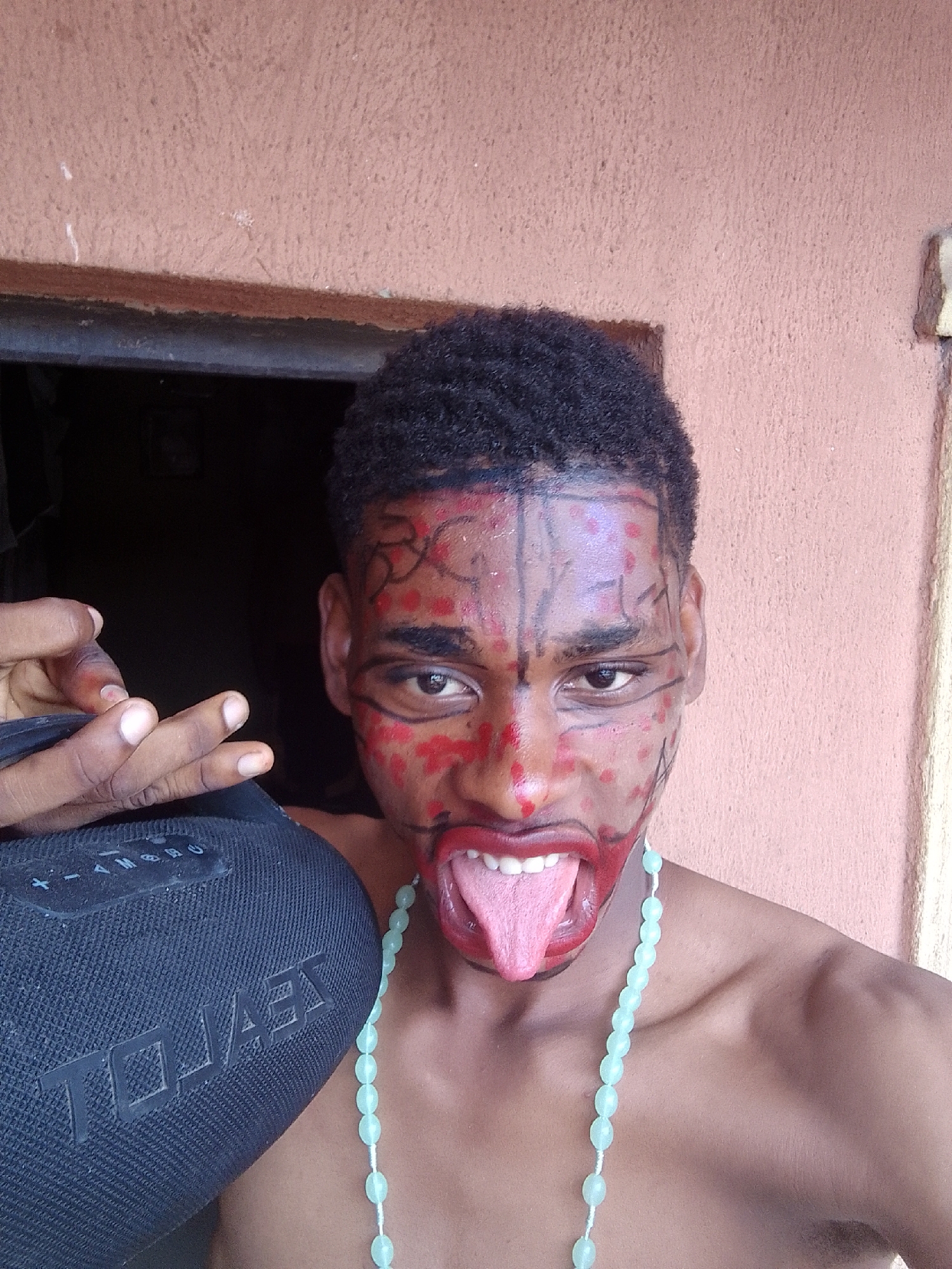 A young person, presumably male, with dark skin and short, curly dark hair, is captured in a close-up, chest-up shot. They are looking directly at the camera with an expressive, energetic pose, featuring wide-open eyes and an extended tongue. Their face is elaborately painted with red dots, concentrated around the nose, cheeks, forehead, and lips, and distinctive black linear designs. Black lines define the eyebrows, underline the eyes, trace down the forehead, and adorn the cheeks and jawline, while the lips are outlined in black and filled with red.
The person appears shirtless and is wearing a light blue/green beaded necklace with oval-shaped beads. They are holding a black cylindrical portable speaker, which is covered in a mesh-like fabric, with their left hand. The brand "JBL" is visible in white letters on the speaker, along with faint power and volume symbols.
The background consists of a textured, light reddish-brown wall to the right and above the subject, suggesting an indoor or sheltered outdoor setting. To the left and behind the person, there is a dark, recessed area, possibly a doorway or opening. The lighting appears natural and bright, illuminating the subject clearly. The overall impression is one of playful self-expression or a costumed appearance.
