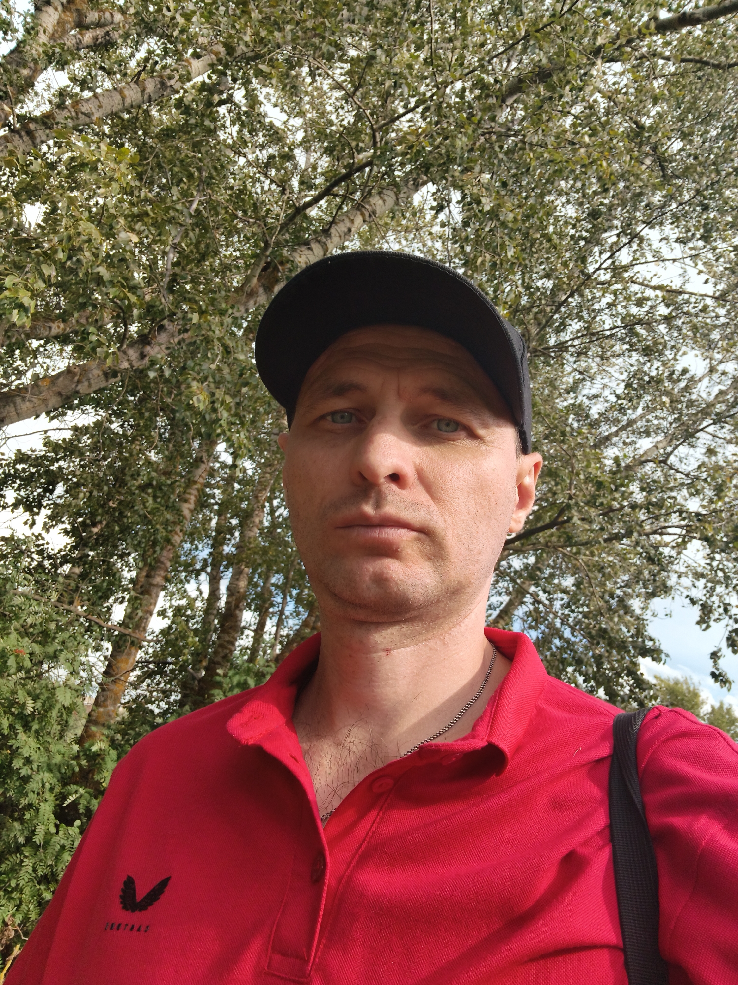 A mid-shot, selfie-style photograph shows a Caucasian male with light skin and blue eyes. He is wearing a black baseball cap and a red collared shirt. A silver chain necklace is visible around his neck. The man is standing outdoors with a backdrop of trees and foliage, suggesting a park or natural area. The lighting indicates it is daytime, possibly overcast. The image is taken from chest-up, focusing on the man's face and upper torso. The background features a mix of green leaves and pale bark from birch trees. The overall impression is a casual outdoor portrait. The user specifically requested the location be "Mokshan, Russia", which cannot be determined from the visual information presented in the image.