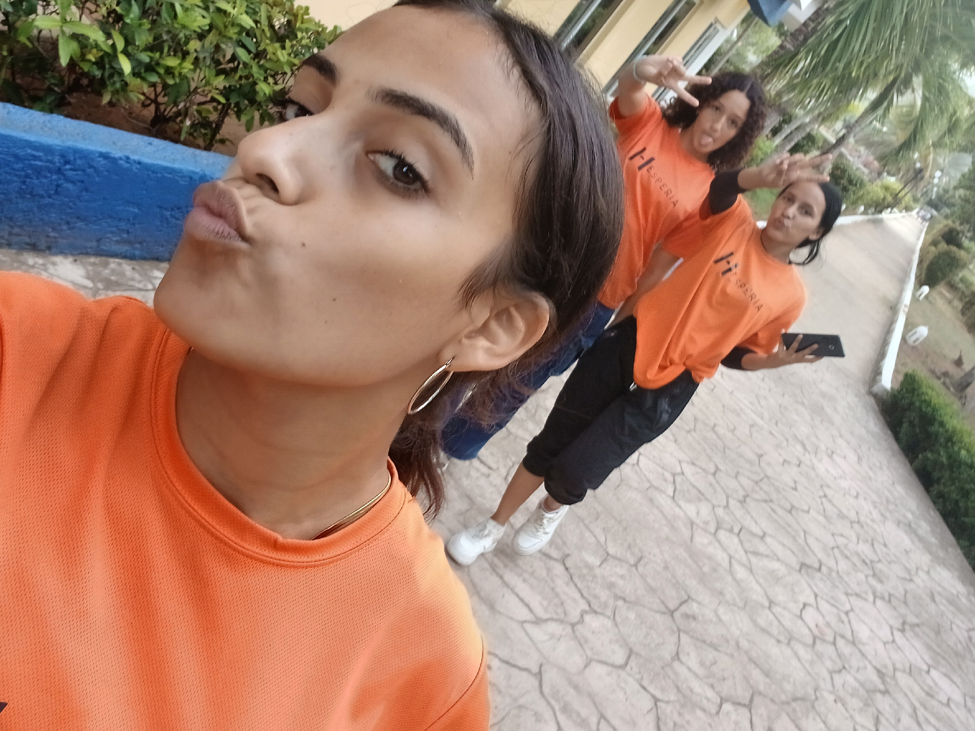The image shows three young women at Playa El Agua, Venezuela. Two of the women are in the background, wearing orange t-shirts and making peace signs. One is holding a smartphone. The third woman is in the foreground, taking a selfie and sticking her tongue out. She is also wearing an orange t-shirt. The ground is paved with a cracked pattern. There are palm trees and green bushes in the background.