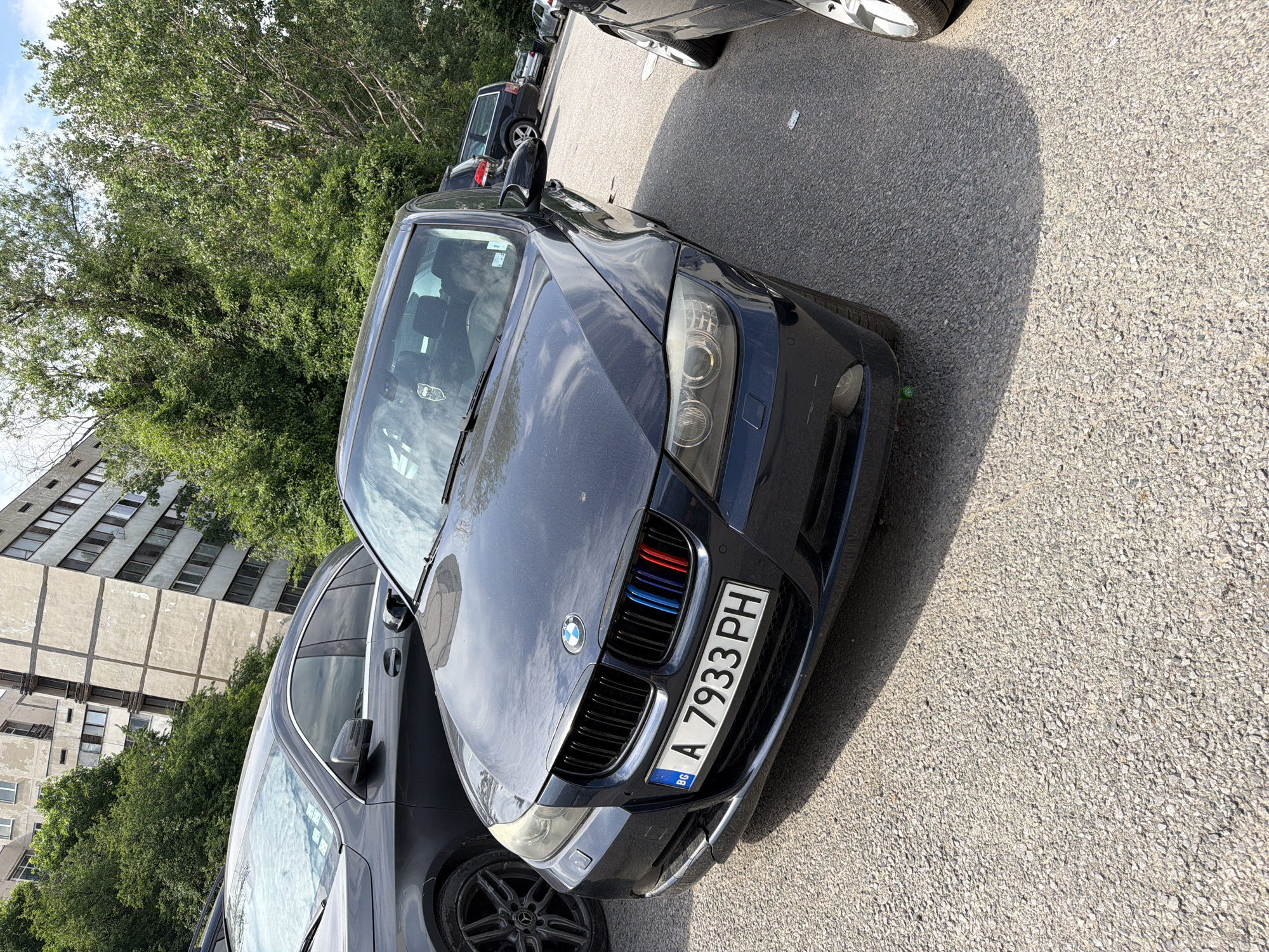 The image displays a dark blue BMW E90 sedan parked in an outdoor asphalt parking lot or street. The vehicle bears a Bulgarian license plate "BG A 7933 PH," with "A" denoting the Sofia region. The BMW's front grille features three colored stripes (blue, light blue, red) on the left side. Its headlights appear aged with some haziness. A green valve cap is visible on the front driver's side wheel.
To the immediate left of the BMW, the rear portion of a dark grey Mercedes-Benz SUV/Crossover is partially visible, featuring a black multi-spoke alloy wheel with the Mercedes-Benz logo. Further in the background, other parked vehicles, including a dark wagon or SUV, are discernible.
The ground is a light grey asphalt surface composed of coarse aggregate, with sunlight casting distinct shadows from the parked cars, indicating bright outdoor conditions.
The background features tall, multi-story buildings, likely residential, with a worn facade characteristic of socialist-era construction. These buildings are partially obscured by dense green foliage from numerous trees and bushes, suggesting a well-vegetated urban environment. The combination of the Bulgarian license plate and the architectural style confirms the location as the city of Sofia, Bulgaria. No people are visible in the scene.