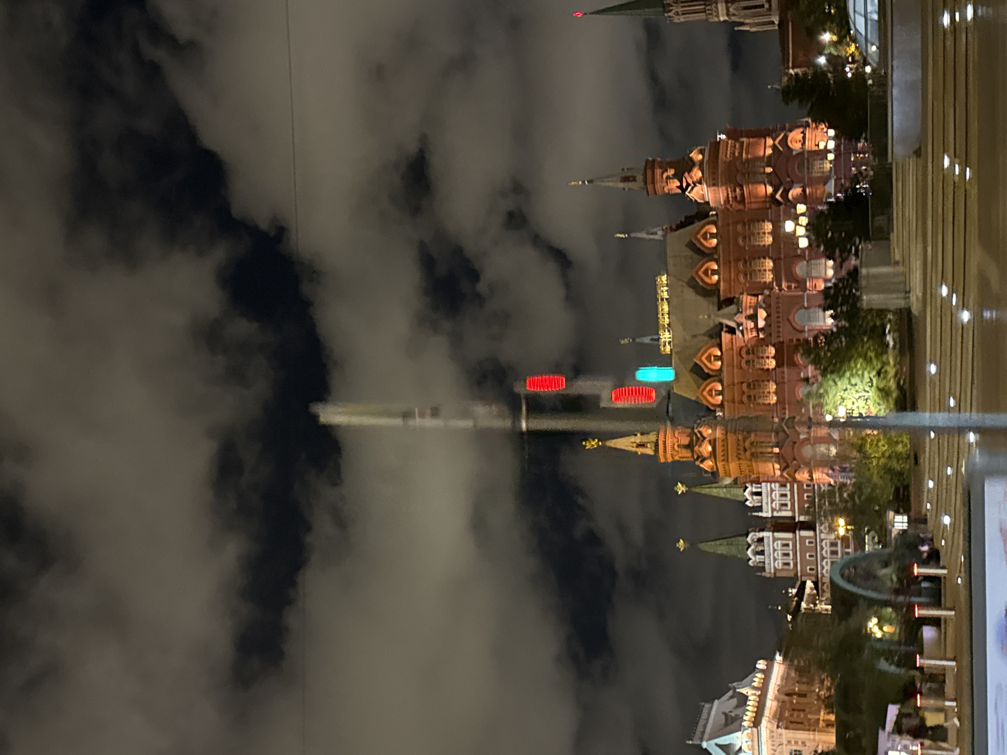 The image captures a nighttime view of Red Square in Moscow, Russia. The scene is dominated by several historic buildings, illuminated by artificial lights. The State Historical Museum, a large red brick building with ornate architecture, is prominently featured in the center of the frame. To its left, the facade of the Kazan Cathedral is partially visible. The background is filled with dark, cloudy skies, with a tall structure, possibly a tower or a light pole, rising vertically and featuring red and green lights, adding a modern element to the historic setting. The ground level shows a well-lit walkway or plaza with some reflective surfaces, suggesting it might be wet. In the foreground, there are some blurred elements, possibly indicating movement, and a few indistinct figures. The overall impression is one of a grand and illuminated historical landmark at night.