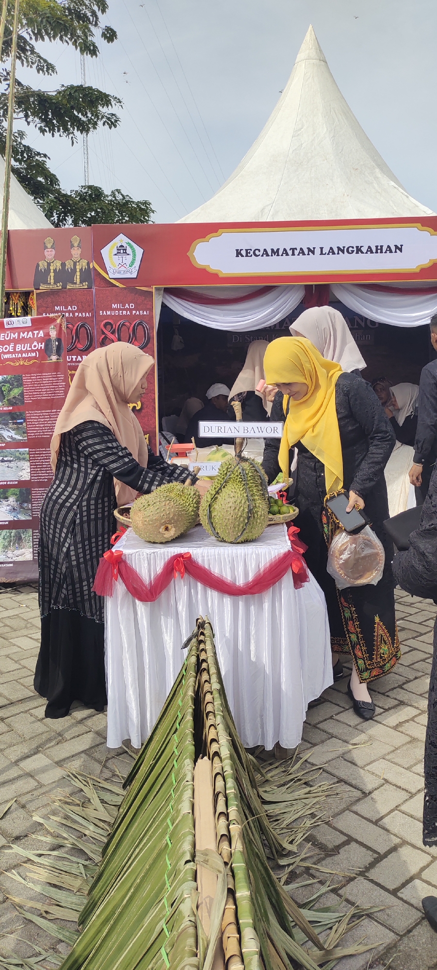 In an outdoor market setting, a table draped with a white cloth and adorned with red ribbon displays two durians and a small basket of limes. A woman in a beige hijab and black and grey checkered dress stands behind the table, reaching towards the durians. Another woman in a bright yellow hijab and black attire with embroidered patterns on the skirt is to the right, holding a mobile phone and a woven plastic bag containing a round, flat object. Behind them, a red tent with white trim is visible, featuring the text "KECAMATAN LANGKAHAN" and some banners with images and text. The ground is paved with interlocking bricks. In the foreground, a triangular structure made of green leaves and bamboo stalks is laid out. The overall scene suggests a local event or festival showcasing regional produce.