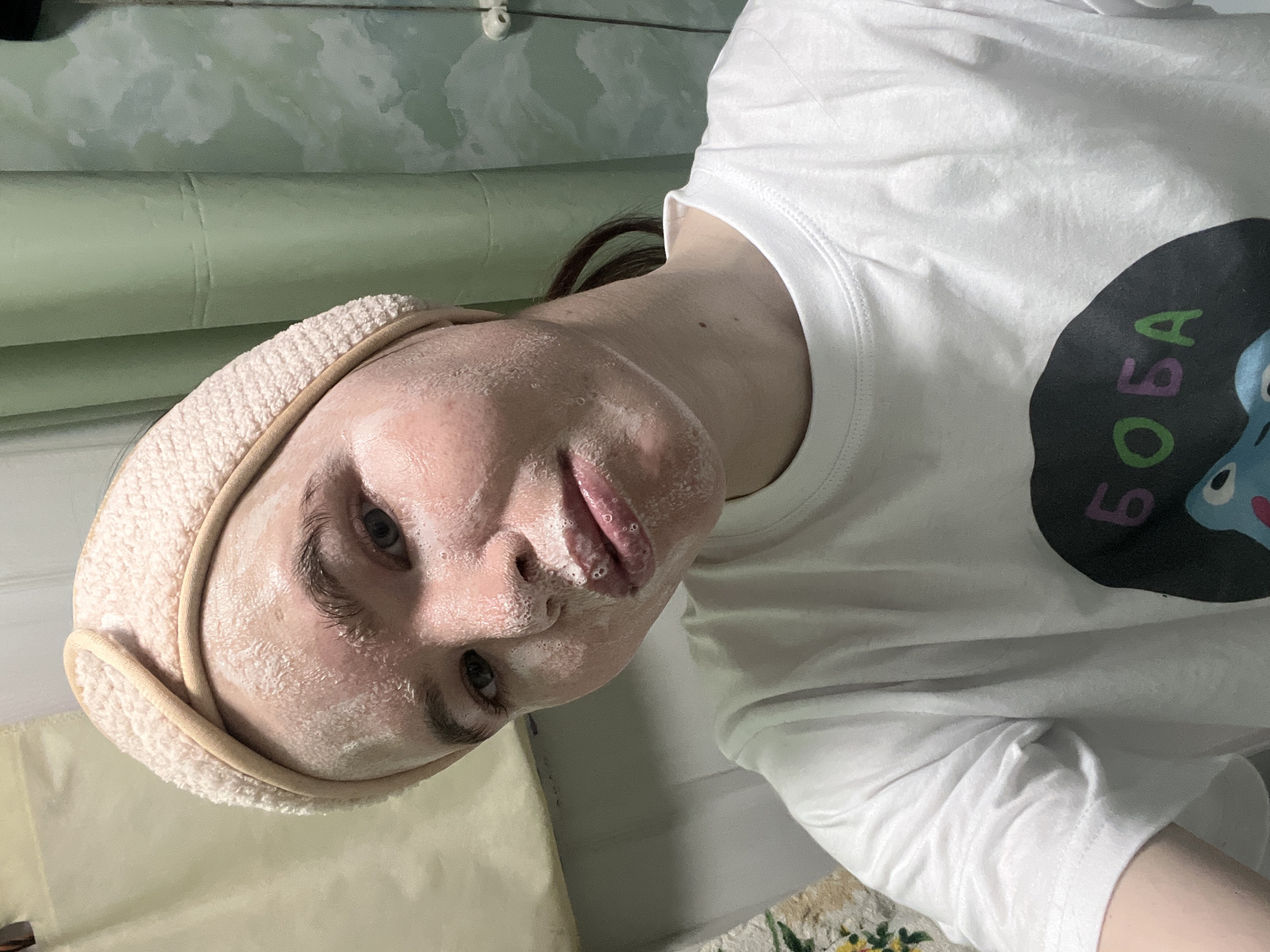 An individual is depicted in a close-up selfie within an indoor setting, identified as a private residence in Chaikovsky, Russia. The subject, appearing to be a young person with fair skin, faces the camera directly. A light-colored, possibly peach-toned, terry cloth headband is worn, securing the hair away from the face. The face, including the forehead, cheeks, nose, and chin, is coated with a white, foamy substance, consistent with a facial cleanser or mask; distinct bubbles are visible, particularly around the lips and nasal area. The individual wears a white, short-sleeved t-shirt with a dark, circular graphic on the front, partially showing the Cyrillic letters "БОБА". The background on the left consists of a plain, light-colored fabric or wall. To the right, a muted green-blue curtain or wall covering is visible, exhibiting lighter patches, possibly from moisture or wear. A small section of a multi-colored floral pattern, likely on a textile item such as a rug or towel, is partially visible in the lower left portion of the frame.