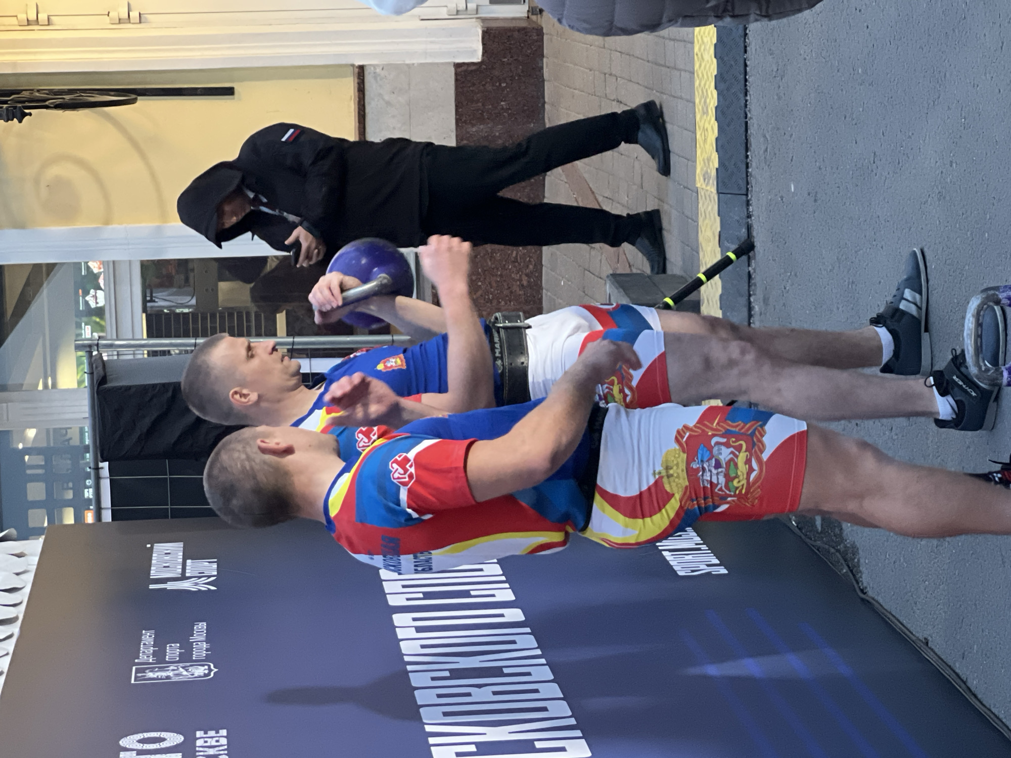 Two men in athletic wear are positioned in front of a banner, with the man in front holding a kettlebell above his head. A person wearing a black hooded jacket and dark pants is in the background. The location is Moscow, Russia, as indicated by the banner which is in Russian.
