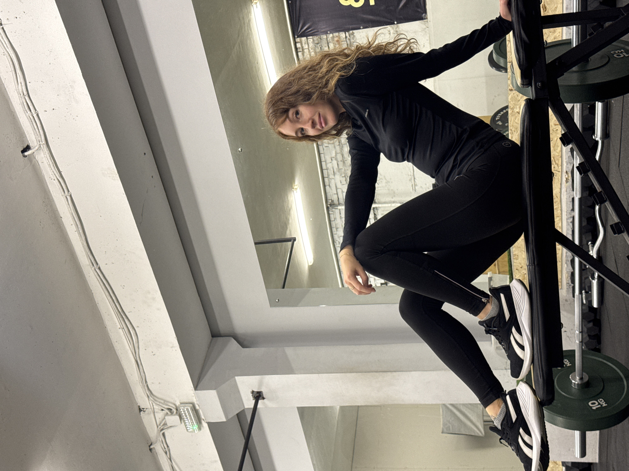 A young woman with curly brown hair is posing in a gym setting. She is wearing a long-sleeved black top, black leggings, and black and white sneakers. She is holding onto a squat rack loaded with weight plates. The gym has a modern industrial aesthetic with exposed concrete and visible pipes. The location is in Chaikovsky, Russia.