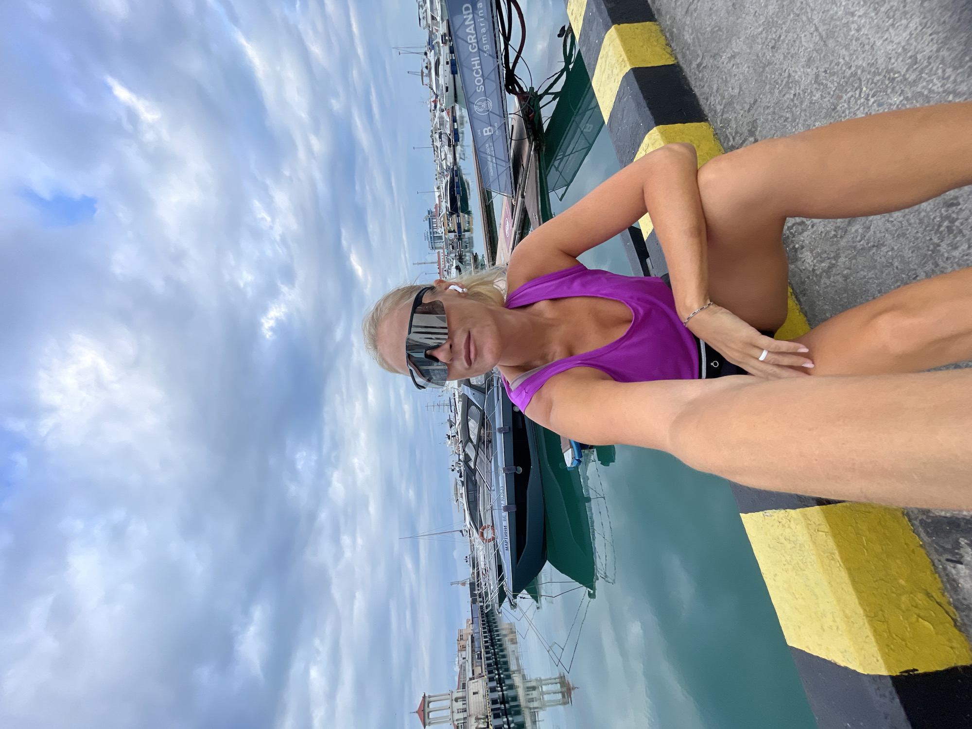 A woman with blonde hair and sunglasses is sitting on a concrete pier next to a marina in Sochi, Russia. She is wearing a purple tank top and black shorts. She is holding a phone in her right hand, appearing to take a selfie. The marina is filled with numerous yachts, and a tower-like structure with a red roof is visible in the background. The water is calm and reflects the cloudy sky and boats. The pier has yellow and black markings on its edge.