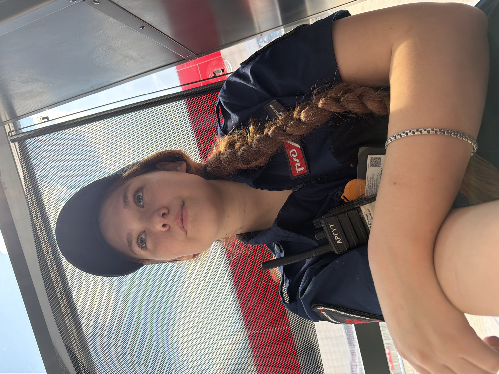 The image shows a young woman wearing a dark blue uniform with a black baseball cap, looking upwards and to the left. She has reddish-brown hair styled in a long braid that falls over her left shoulder. On her uniform, there are patches and a radio clipped to the chest. A silver bracelet is visible on her left wrist. The background suggests an outdoor setting with a bright sky and some red and gray structures, possibly part of a transportation hub or industrial area. The overall lighting indicates it is daytime. There are no identifiable landmarks that specifically place the scene in Rostov-on-Don, Russia.