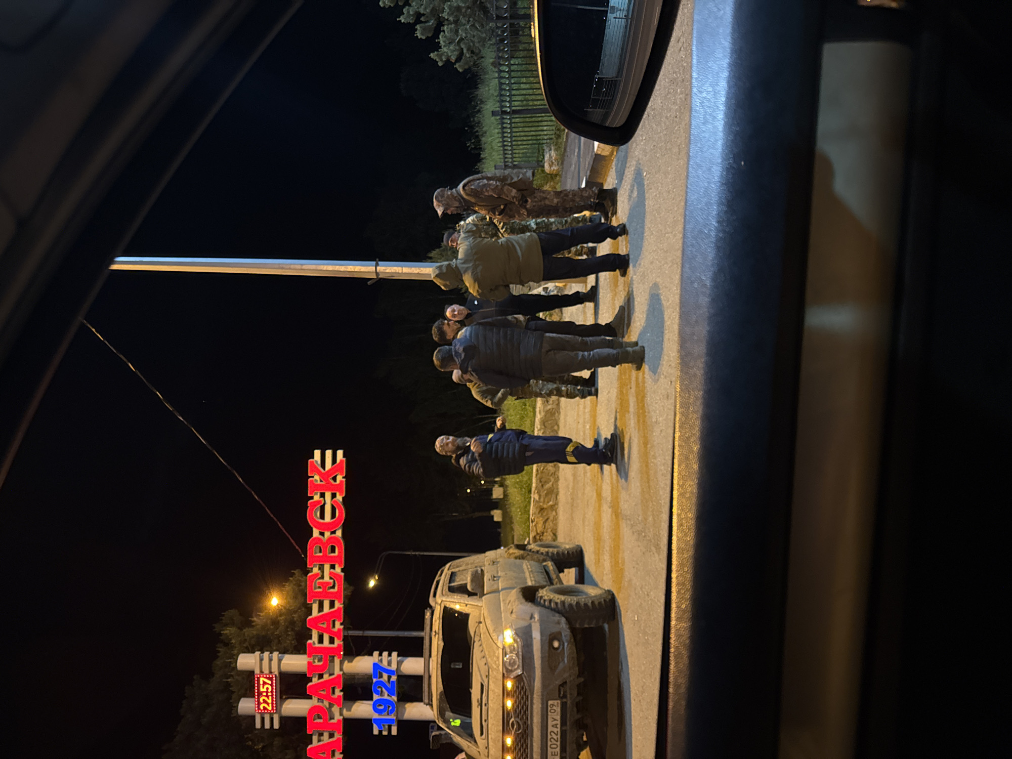 At night in Karachayevsk, Russia, a group of about seven people, some in military-style clothing, stand on a road next to a military-style vehicle. The illuminated "АРАЧАЕВСК" sign is visible, along with a digital clock displaying "22:57" and "1927." The photograph is taken from inside a vehicle.