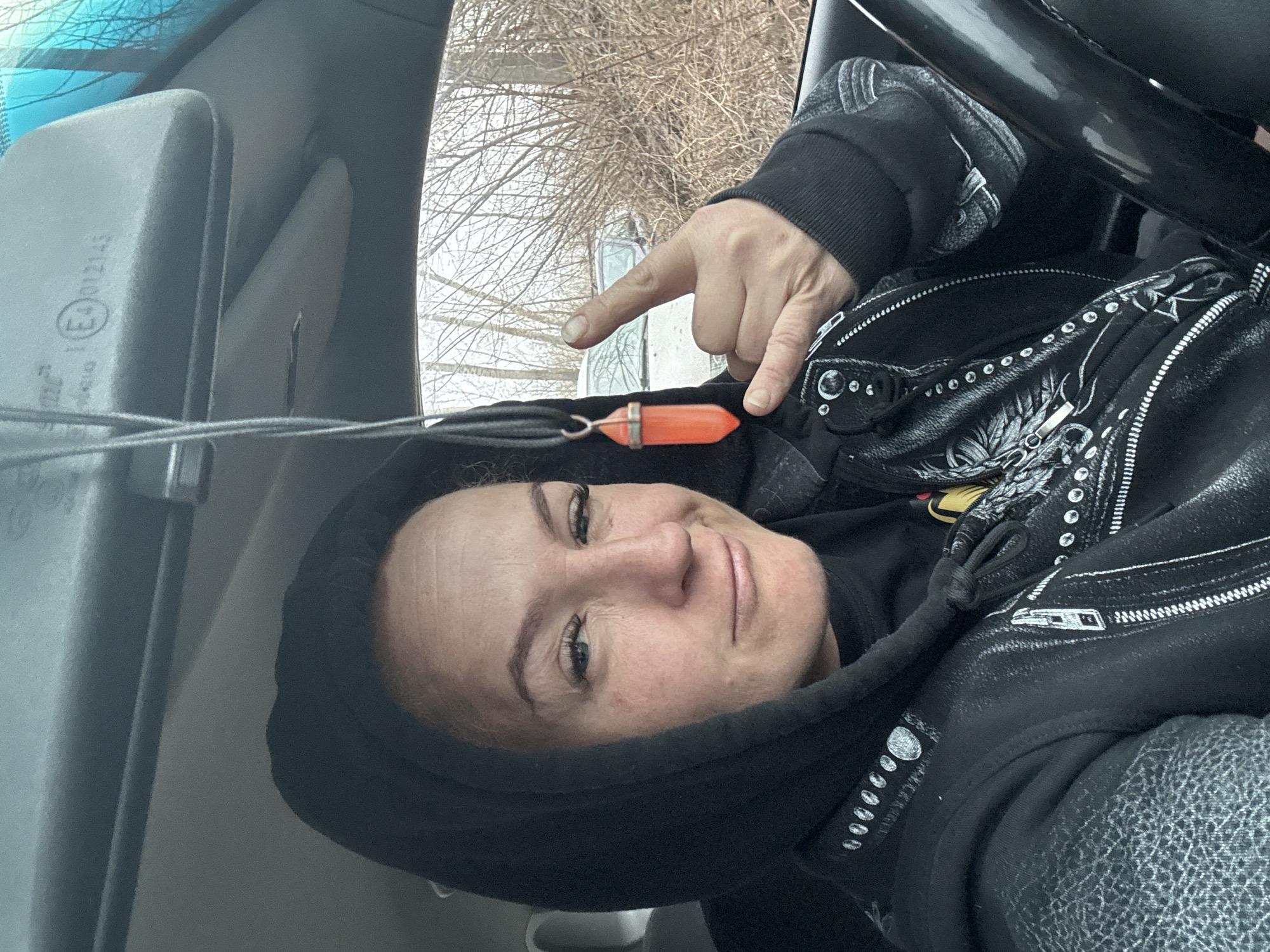 A woman is in a vehicle. The woman is wearing a black hooded jacket with decorative white stitching, including moon phases. She is wearing a pendant on a string. She is gesturing with her index finger toward the pendant. The interior of the vehicle is visible, including the steering wheel and the rearview mirror. The exterior visible through the windows appears to be an area with bare trees. The location is unidentified but could be Chaikovsky, Russia.