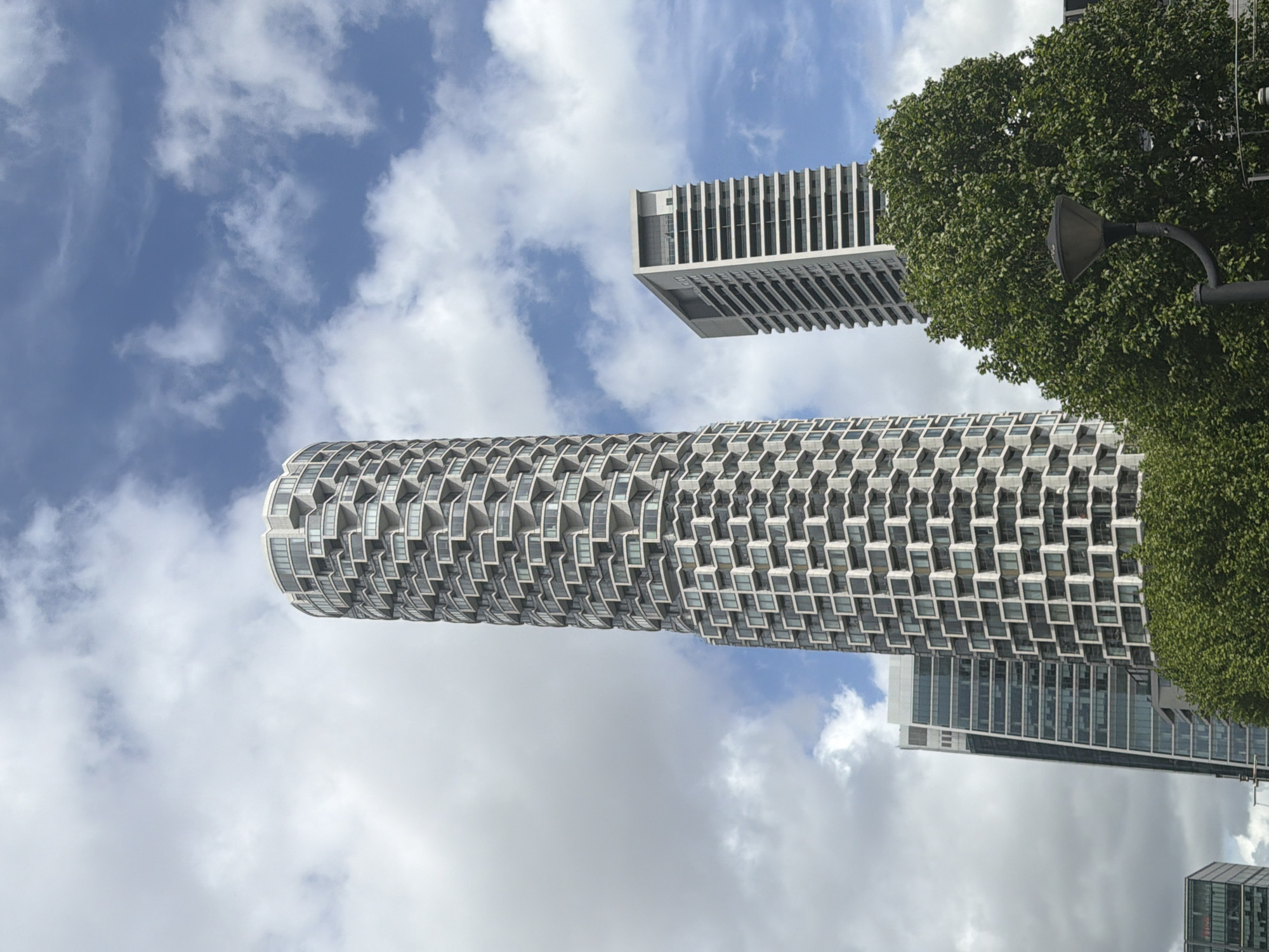 The image shows an upward-angled view of modern architecture in London, United Kingdom. The cylindrical structure of One Park Drive, known for its distinctive honeycomb-like facade of apartments, is a prominent feature. Situated near the smaller, square-shaped building of Newfoundland Quay. Clouds fill the blue sky background, with a leafy tree on the right of the frame near a light post.