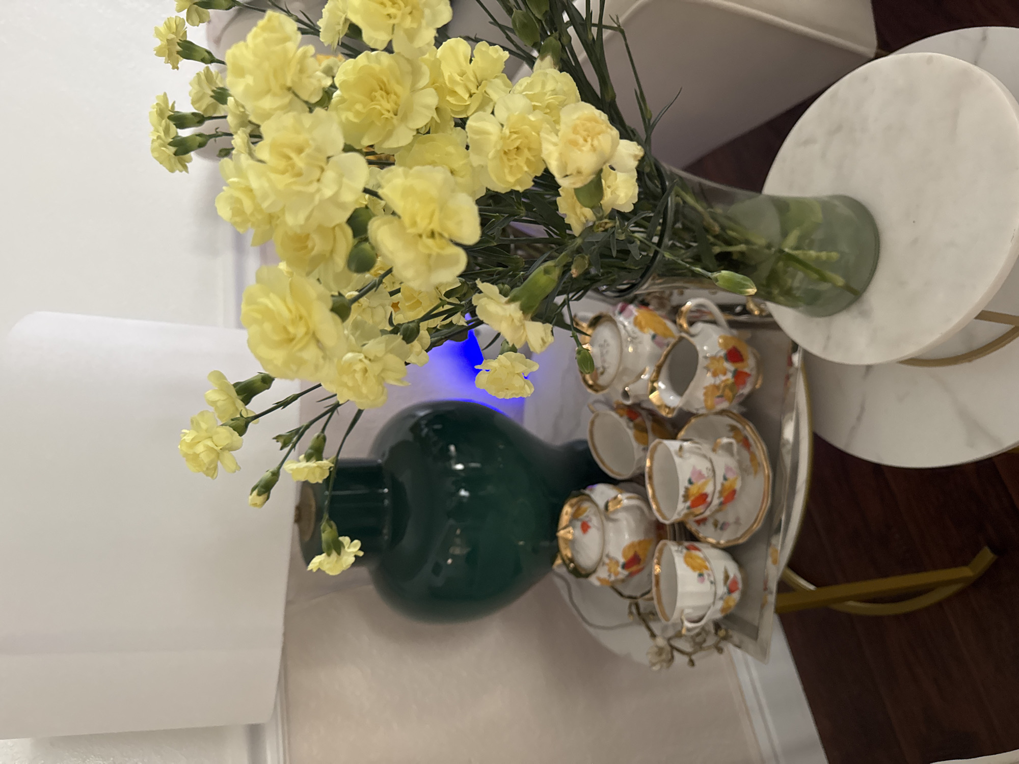 In Clovis, California, a still life photograph captures a domestic setting. A white lampshade dominates the left side of the frame, partially obscuring a tall, emerald green ceramic vase. The vase is situated on a dark wood floor, near a white textured wall. To the right of the vase, a clear glass vase holds a vibrant bouquet of yellow carnations, their delicate petals filling the upper right quadrant of the image.
In front of the green vase, on a silver tray with gold trim, rests a miniature porcelain tea set. The set includes a lidded sugar bowl, a creamer, and several small teacups and saucers. Each piece is adorned with a floral pattern featuring orange, yellow, and pink blossoms, accented with gold trim. The tea set is arranged artfully on the tray, suggesting a decorative rather than functional purpose.
The tray is supported by a slender, gold-colored metal stand. To the right of this stand is a small, round white side table with a marble-like finish. The overall lighting suggests an indoor environment, possibly a living room or dining area, with soft ambient light contributing to a serene atmosphere. There are no people or animals visible in the scene.