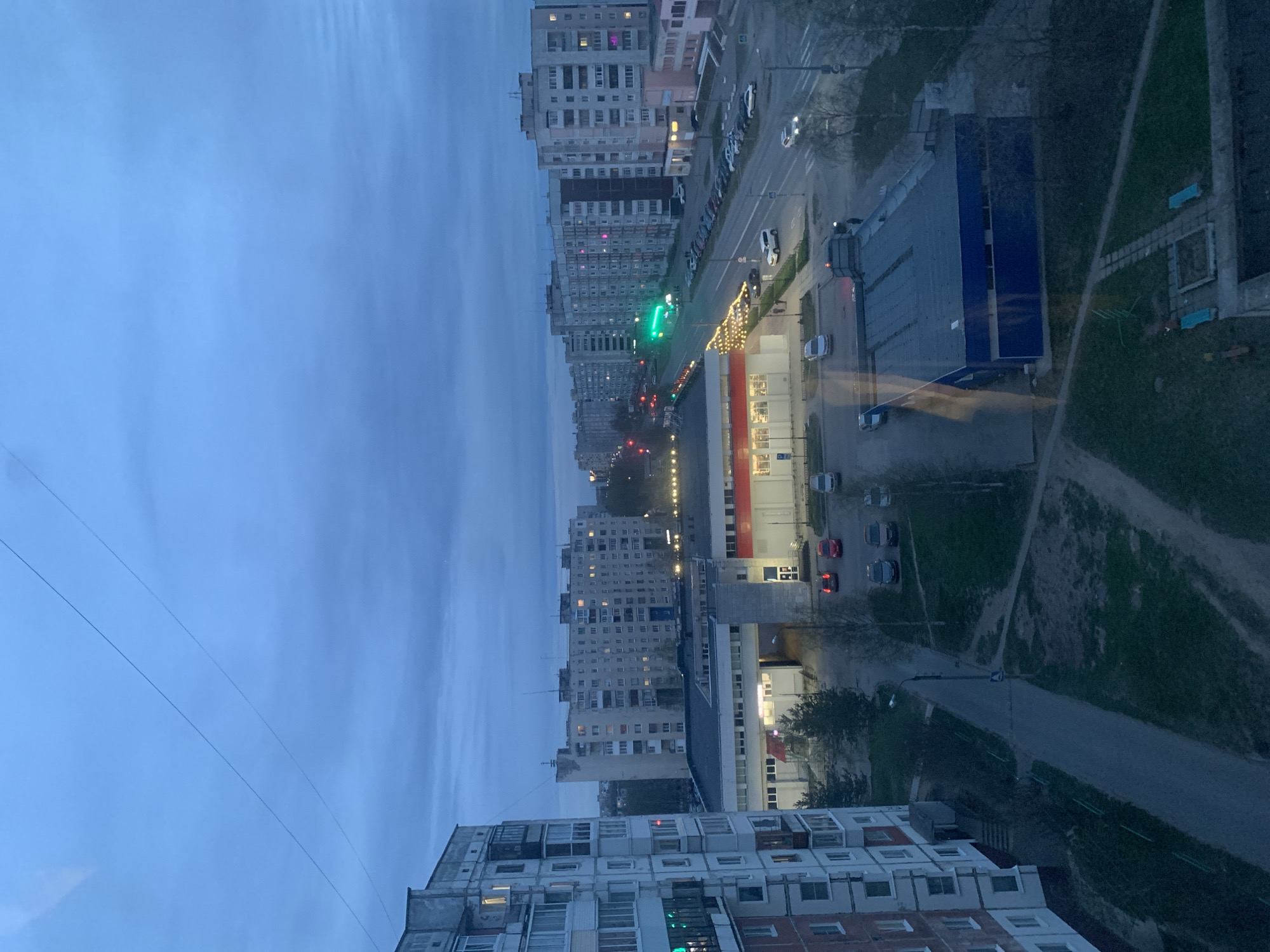 The image presents an elevated view of a residential area in Severodvinsk, Russia, likely taken from an upper floor window. The buildings are mostly high-rise apartment blocks, typical of Soviet-era architecture. A main street runs between the buildings, with traffic lights indicating a green signal. Several vehicles are visible on the road and parked along the roadside. A long building with a blue roof and a red and white facade, likely a shopping center or commercial building, is also present. Adjacent to it is a walkway or path flanked by greenery and trees. The sky is overcast, suggesting dusk or dawn. The overall impression is of a typical urban landscape in a northern Russian city.
