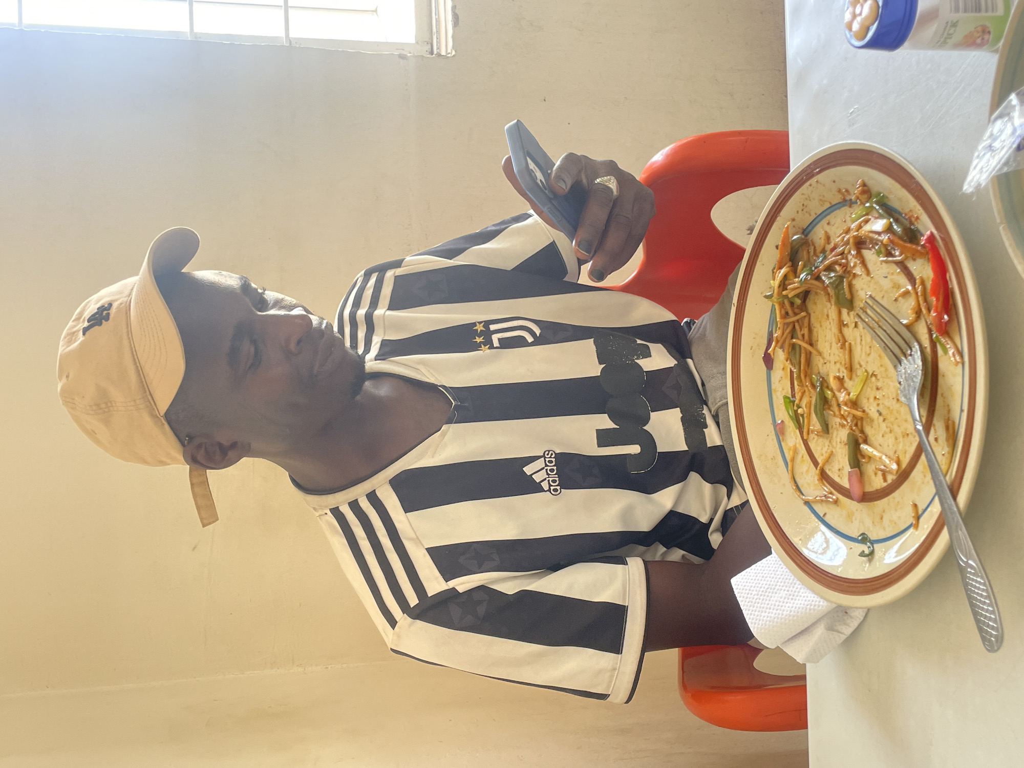 A man wearing a baseball cap and a Juventus soccer jersey is sitting at a table, looking at a smartphone in his hand. On the table is a plate with the remnants of a meal, mostly noodles and vegetables, with a fork resting on it. A napkin is placed to the side of the plate. The location is Jos, Nigeria. The environment appears to be a simple, possibly indoor, setting with a window in the background.