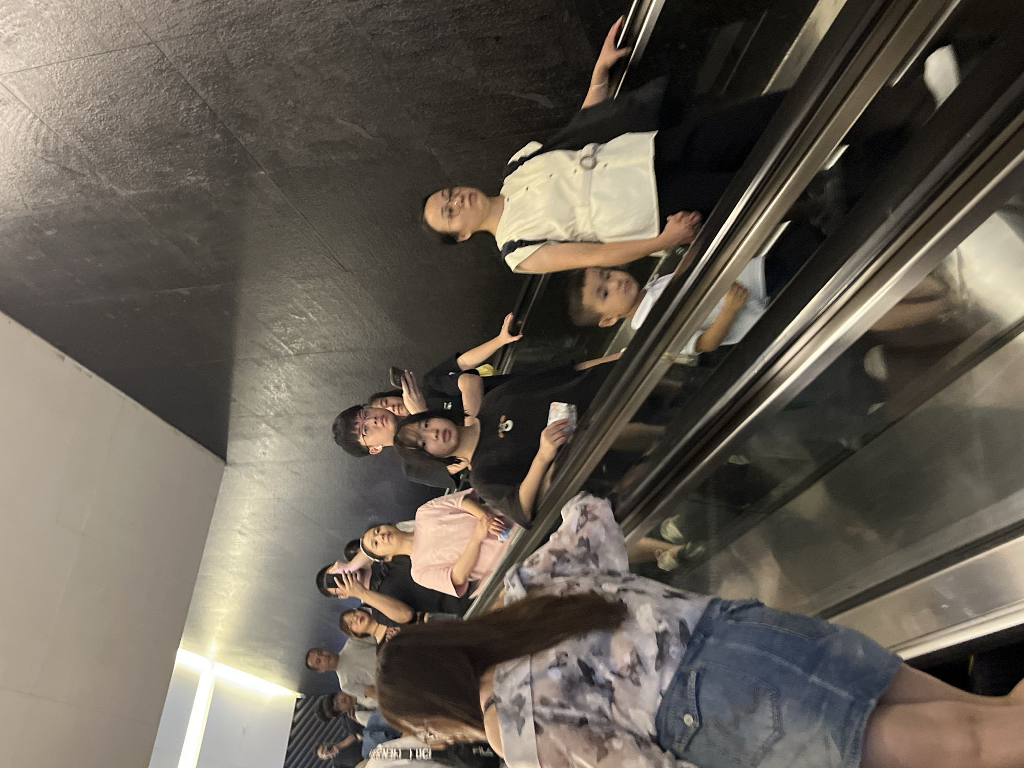In Haizhu District, China, a group of people are ascending an escalator, their reflections visible in the metallic handrails. The escalator's dark gray, textured walls suggest an indoor, likely urban, environment. Several individuals are looking at their phones, while others engage with their companions. A young boy stands close to an adult woman, his hand potentially held. The scene is illuminated by overhead lighting, casting soft shadows. The people are dressed in casual attire, indicative of everyday urban life.