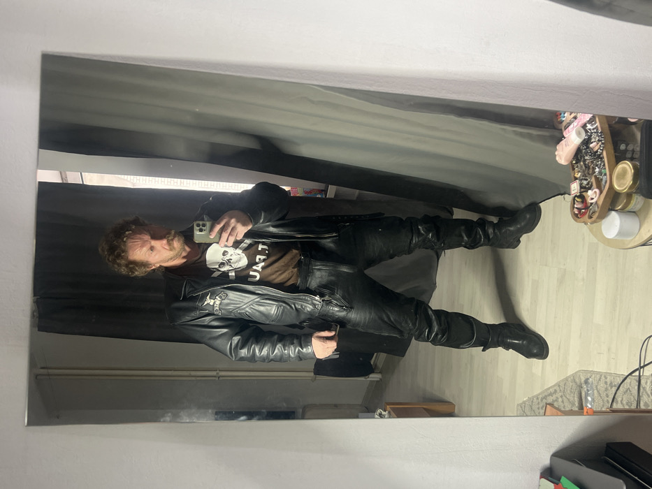 The image displays a full-body mirror selfie of an adult male taken indoors. The precise location within the city of Saint Petersburg, Russia, cannot be determined from the visual evidence.
The subject is a male, appearing middle-aged, with curly light brown hair, a mustache, and a short beard. He maintains a serious expression, looking directly into a light-colored smartphone held in his raised right hand, which captures his reflection. He is dressed in a brown t-shirt featuring a white skull and crossbones graphic on the chest, partially obscured text visible below. Over the t-shirt, he wears an open black leather motorcycle jacket with zipper details and a patch on the left chest. He also wears black leather pants and black leather boots, one of which shows a visible buckle strap. His stance is upright with legs slightly apart.
The reflected background behind the subject reveals a light-colored laminate or wood floor and light grey walls. A dark grey curtain is drawn behind the subject, with a vertical strip of light on its right edge, suggesting a window or light source. On the far left of the reflected background, a light-colored radiator or pipe runs vertically, with an indistinct dark object partially visible below it.
In the actual room, visible in the foreground outside the mirror's frame, a round wooden tray containing various items such as jewelry and small containers, including a pink, textured object, is situated on a cylindrical white stand in the bottom right corner. In the bottom left, a dark electronic device and associated cables are partially visible on the floor.